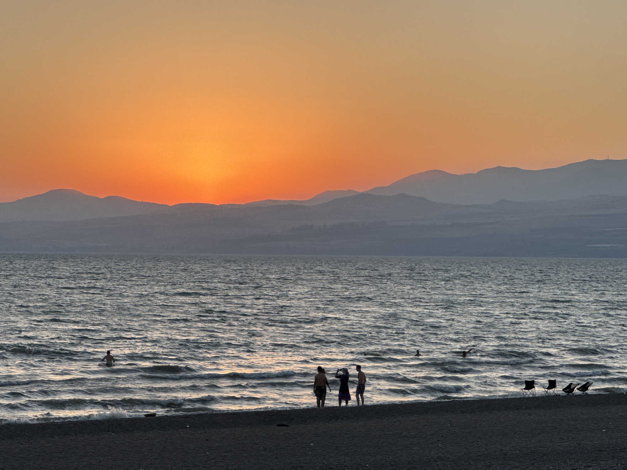 A wide shot captures a serene sunset over the Sea of Galilee, located in the Golan Regional Council, Israel. The sky transitions from a vibrant orange near the horizon, where the sun is partially obscured by mountains, to a soft gradient of peach and yellow. Silhouetted mountains form a hazy backdrop against the colorful sky.
The foreground of the image is dominated by the calm, rippling waters of the sea. Gentle waves lap at a dark, pebbly beach. Several people are visible in and near the water. A lone swimmer is in the middle distance, appearing to float. Closer to shore, three figures stand together on the beach, one of whom appears to be holding a camera. Further out in the water, two other individuals are swimming or wading. On the beach, three empty camping chairs are set up, facing the water, suggesting a place of relaxation.
The overall atmosphere is peaceful and tranquil, characteristic of a late afternoon or early evening at the seaside.