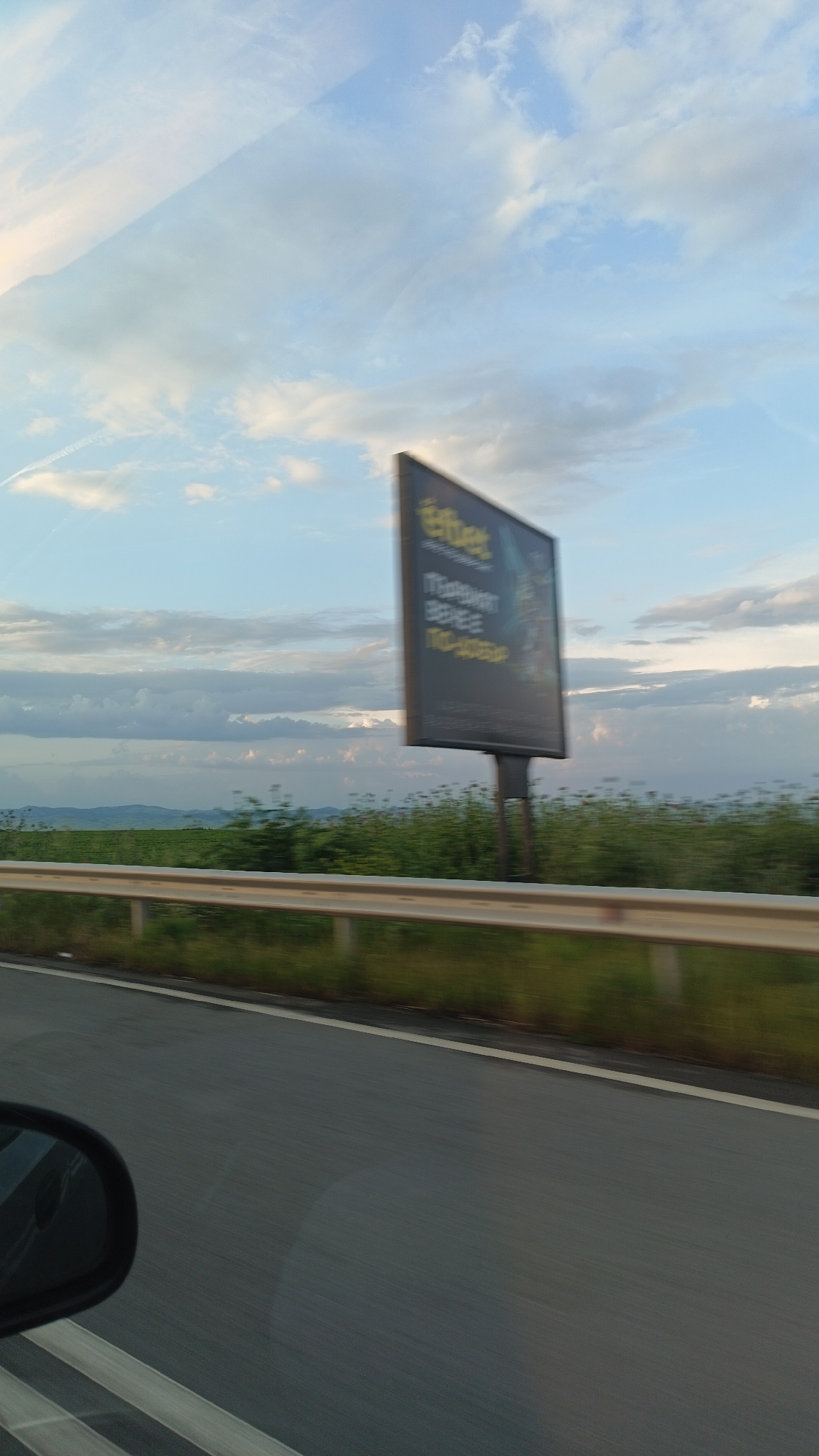The image shows a view from a moving vehicle on a highway. The road surface is dark gray, with white lane markings. A metal guardrail runs along the right side of the road, separating the roadway from a grassy area. Beyond the grass, a field stretches towards the horizon, with hints of hills or mountains in the distance. A large billboard stands in the field, its advertisement partially blurred due to the motion. The sky is blue with scattered clouds, and contrails are visible. A portion of the car's side mirror is visible in the lower left corner of the image. Given the billboard's branding and the landscape, the location is likely near Burgas, Bulgaria, but more specific location details are difficult to ascertain.
