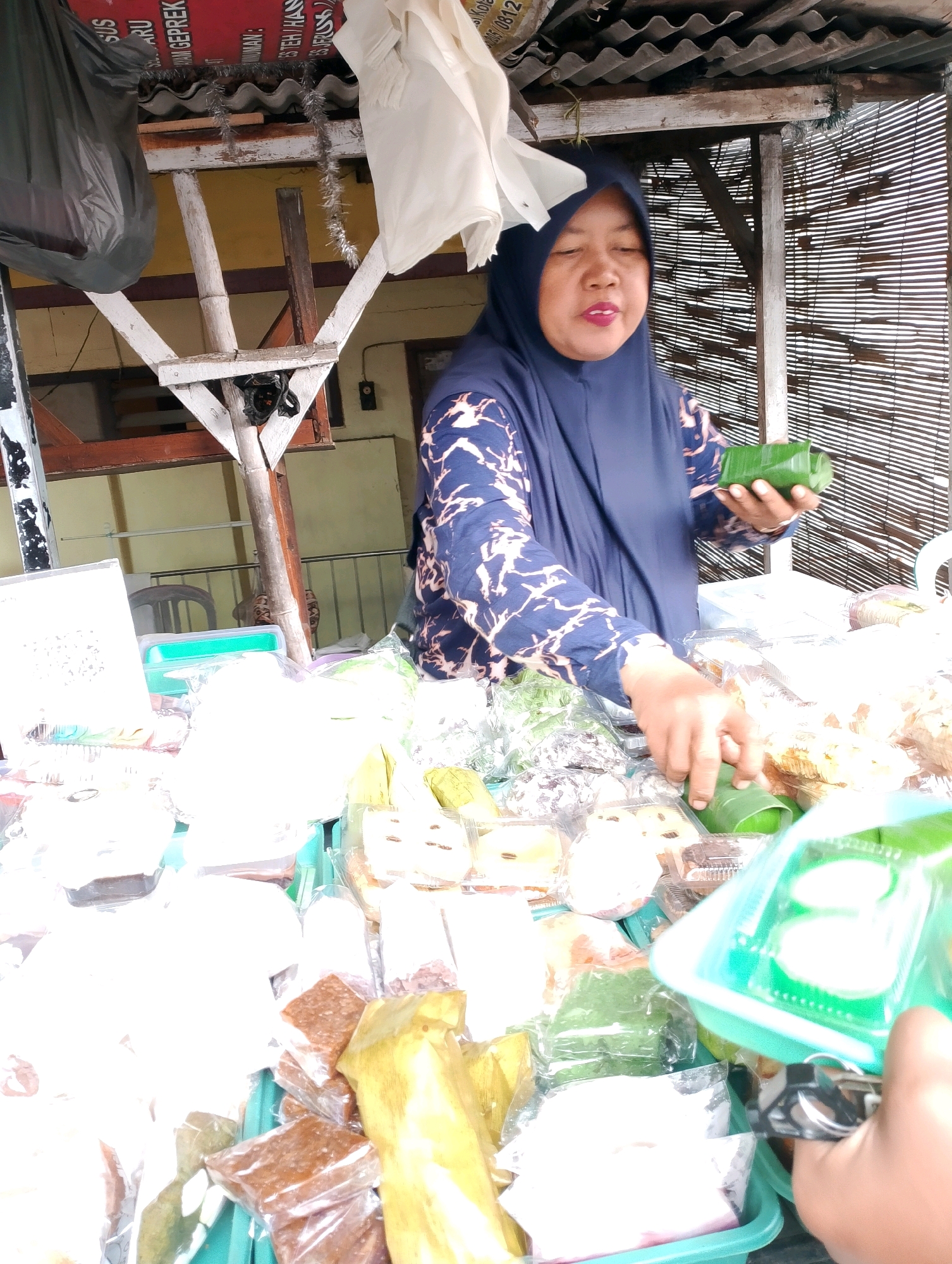An outdoor market stall in Probolinggo, Indonesia, displays a variety of snacks. A woman wearing a dark blue hijab and a patterned long-sleeved shirt is seen behind the stall. She is holding a snack wrapped in green leaves in her right hand and her left hand is reaching for another similar snack. The stall is filled with an assortment of packaged food items, including some wrapped in plastic and some in green leaf packaging. The background shows a corrugated metal roof, bamboo blinds, and some signage that is partially visible. A black plastic bag hangs from the stall structure on the left. The scene appears to be a busy marketplace.