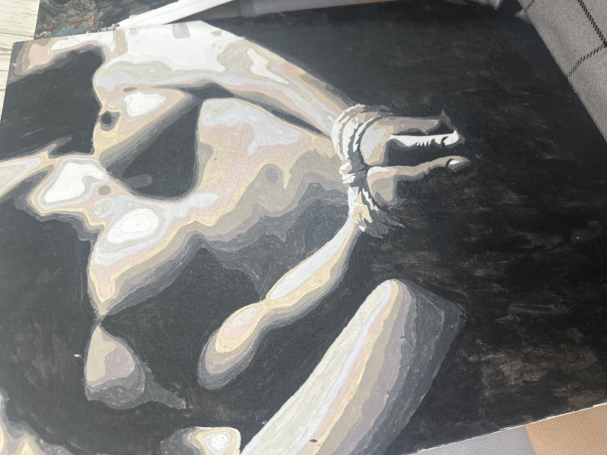 A canvas painting depicts a figure with bound hands against a dark background. The figure is rendered in grayscale tones with distinct color-blocked sections, suggesting a paint-by-numbers style or an artistic simplification of form. The subject appears nude or partially nude, with emphasis on the torso, arms, and hands. Ropes or bindings are tightly wound around the wrists. The background is primarily a deep, murky black, providing stark contrast to the lighter tones of the figure. The painting has visible canvas texture, and the edges of the canvas are visible in the image. No information indicates the location as Высокий, Russia.
