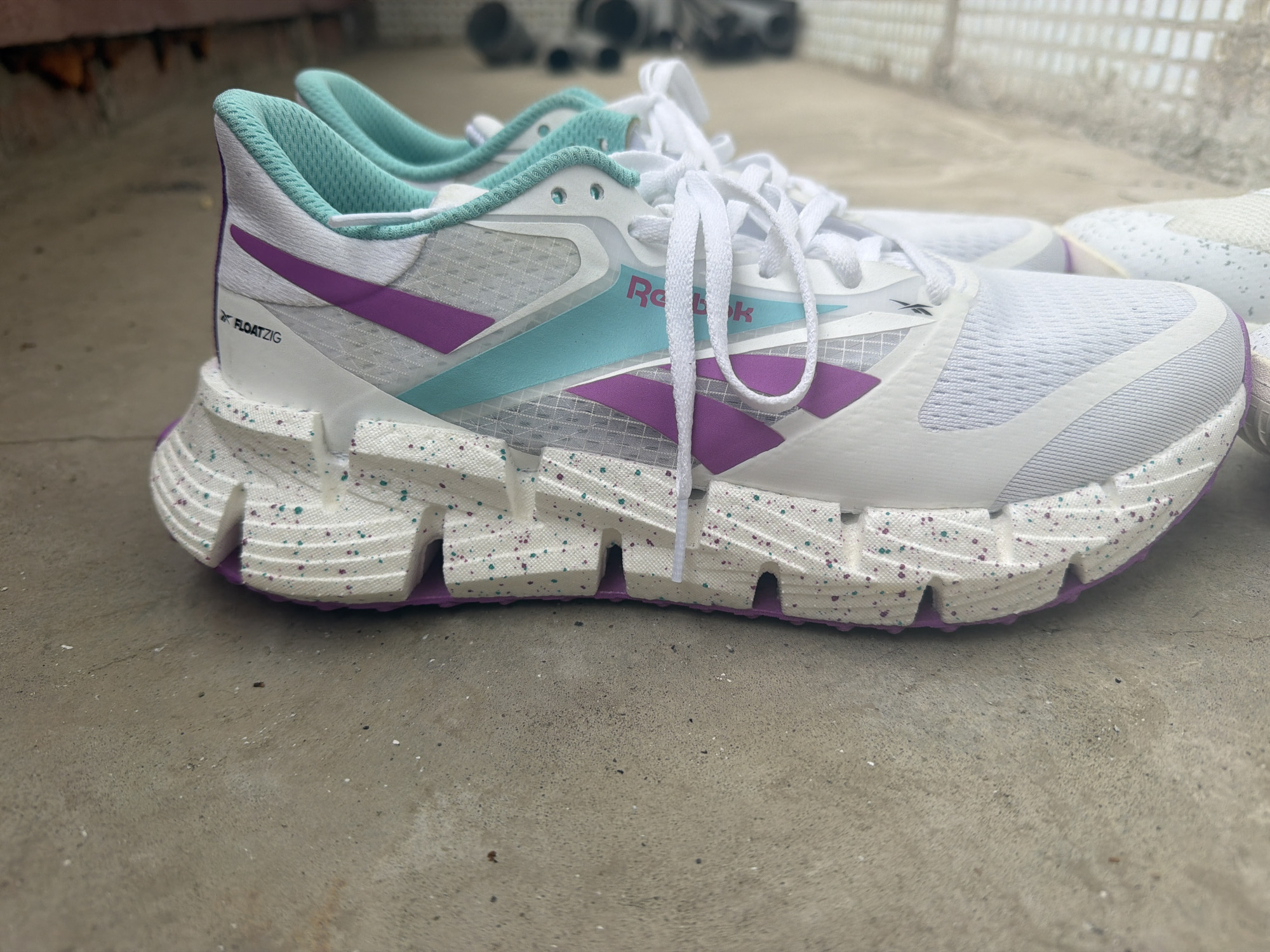 A close-up shot of a pair of Reebok FloatZig running shoes on a concrete surface. The shoes are predominantly white with purple and teal accents. The soles are speckled with blue and purple dots. The laces are white and tied. The location is not identified as Chaikovsky, Russia. Other shoes are visible in the background.