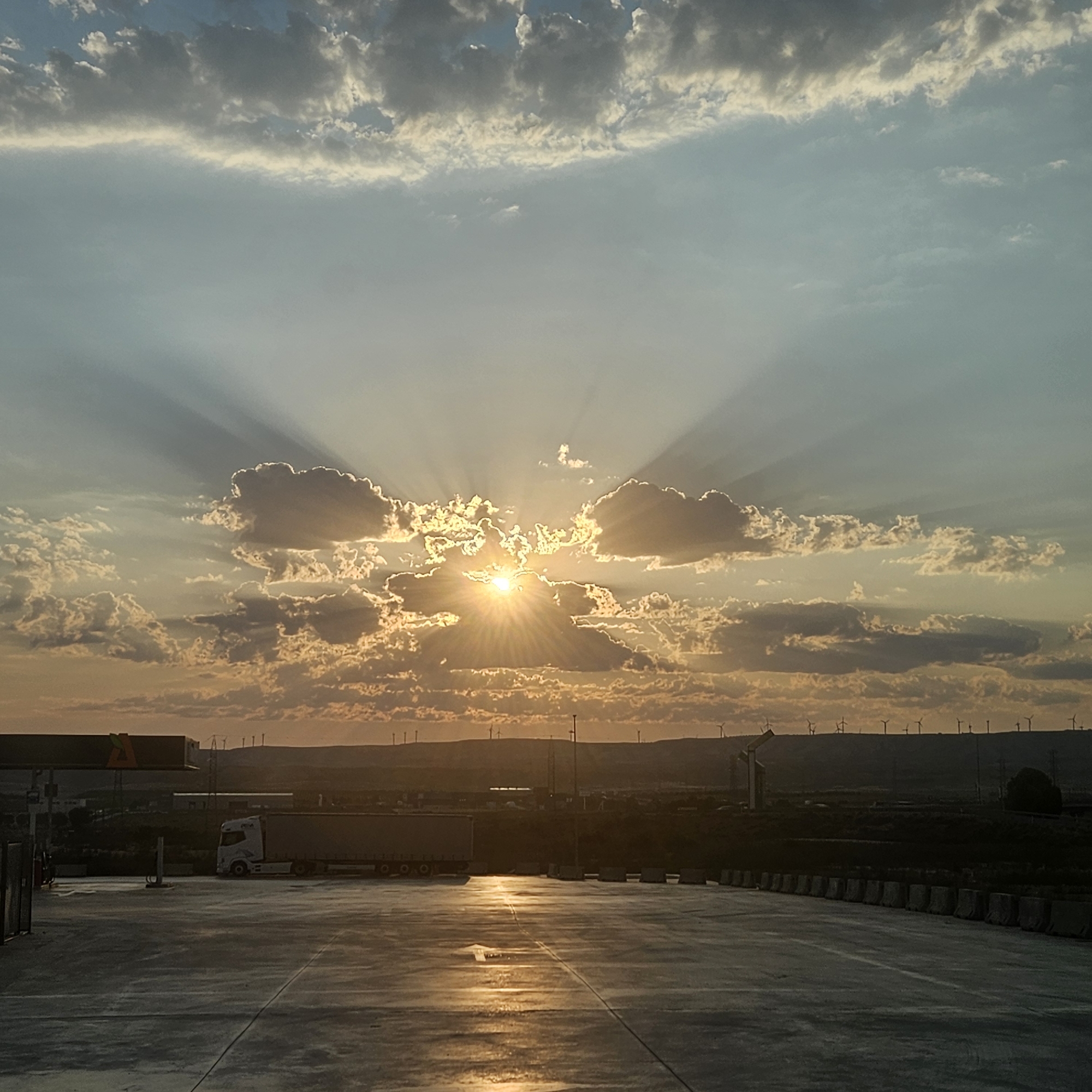 The image captures a sunset over a rest stop or truck stop facility located on the outskirts of Épila, Spain. The sky is filled with dramatic clouds, with the setting sun prominently visible behind a cluster of them, casting crepuscular rays. The foreground consists of a paved area, likely a parking lot, reflecting the warm tones of the sunset. To the left, a white truck is parked near a gas station canopy, its trailer covered. In the background, rolling hills are dotted with numerous wind turbines, characteristic of the Aragonese landscape. The overall impression is one of a quiet, transitional moment at the end of the day in a semi-industrialized rural setting.