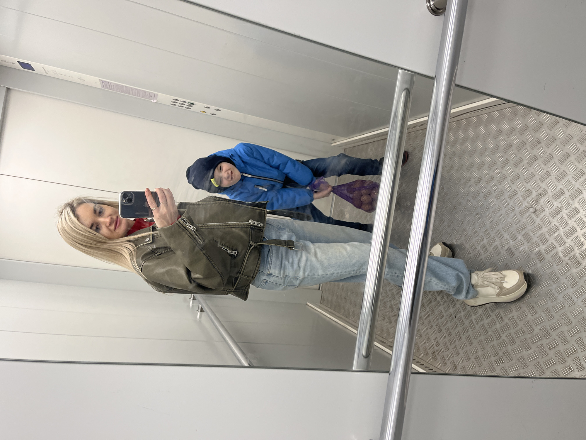 A woman is taking a selfie in an elevator in Chaikovsky, Russia. The reflection reveals a young boy in a blue jacket and dark hat, holding a bag of purple potatoes. The woman wears a green leather jacket, jeans, and white sneakers. The elevator has a metal floor and a white interior.