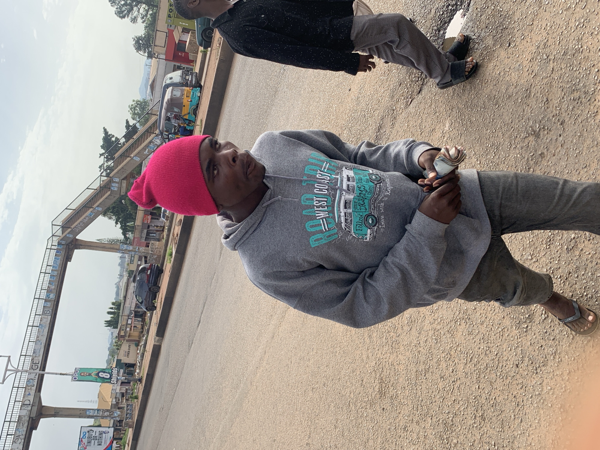 In Gura Top, Nigeria, a young Black male wearing a bright pink beanie and a grey hooded sweatshirt with "WEST COAST" and a graphic of a camper van printed on it, is walking towards the right of the frame, holding a wad of cash in his right hand. His facial expression is one of mild surprise or exertion. He is wearing dark-colored pants and flip-flops. Behind him and to his right, another Black male is walking away from the camera, wearing a dark patterned shirt and light-colored pants, also in flip-flops. They are on a dirt and asphalt surface, possibly a road or pathway. In the background, a concrete pedestrian bridge spans overhead, adorned with graffiti. Various businesses and buildings are visible along the side of the road, including signs for "OPay Shields" and "CYBER CAFE". Several vehicles, including a van and what appears to be a tuk-tuk, are also present in the background. The sky is overcast.
