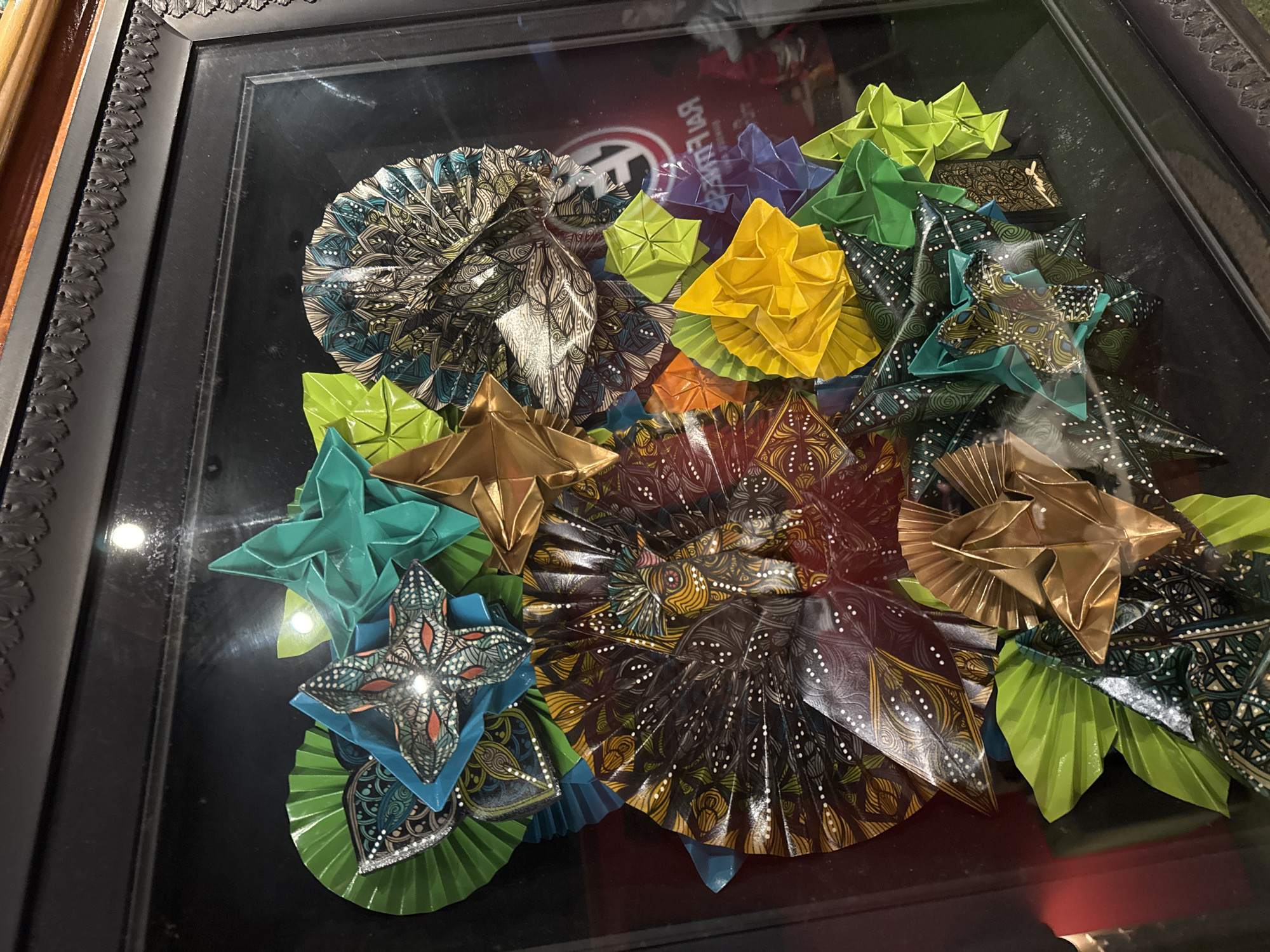 The image shows a framed artwork composed of various origami creations. The artwork is displayed within a black frame with ornate detailing along the top. The origami pieces are arranged in a cluster and feature a variety of colors and patterns. Some of the origami appear to be fan-shaped or star-shaped, and many are made from patterned paper, showcasing intricate designs in colors like teal, gold, green, yellow, purple, and dark brown with white accents. The background within the frame is black and shows reflections, indicating the artwork is behind glass. A logo, possibly for "Raw Fruits," is faintly visible through the glass in the lower right quadrant of the image. The overall impression is of a decorative and intricate piece of art.