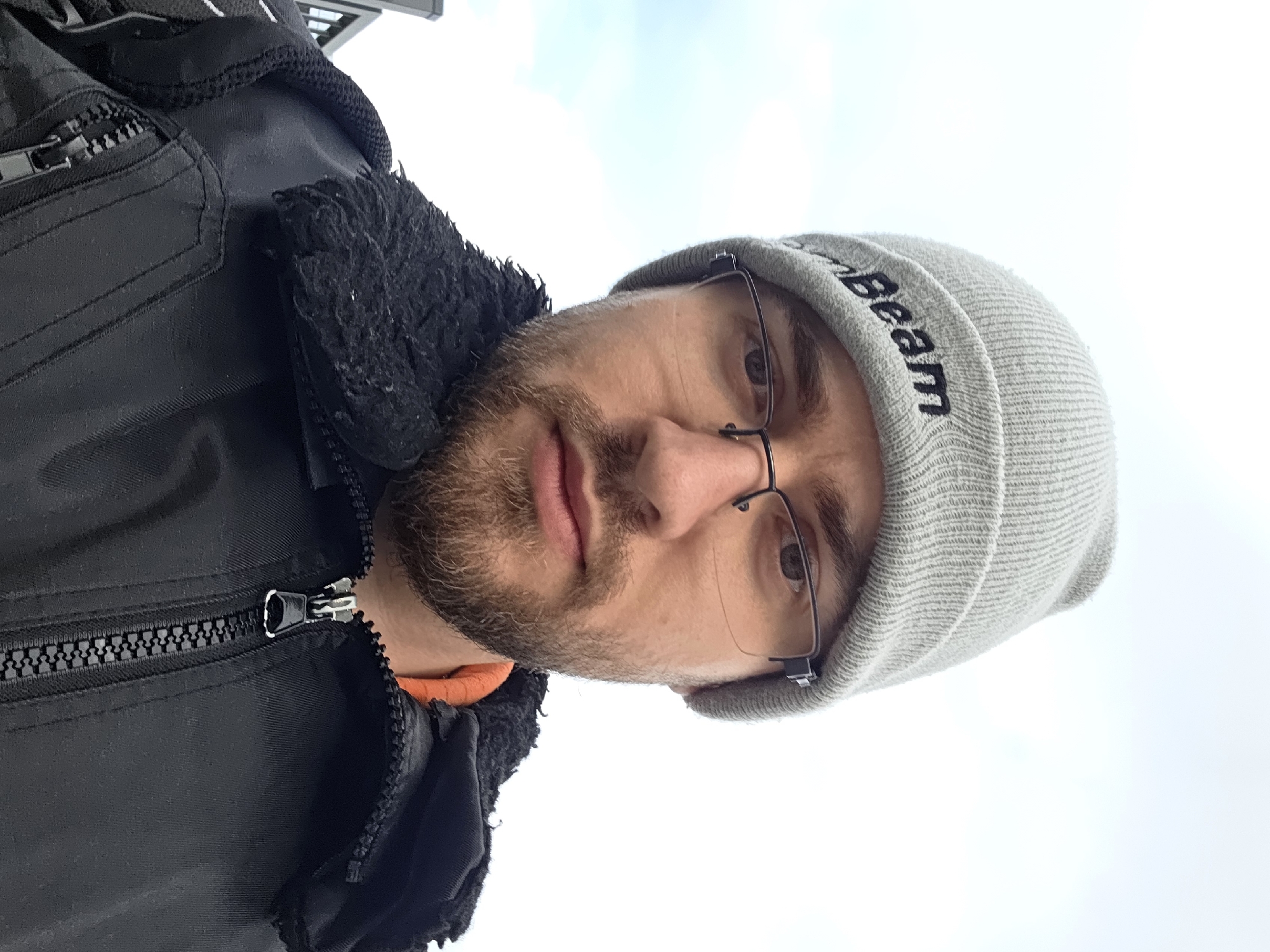 A head-and-shoulder shot of a man with a beard wearing a grey beanie and glasses, taken outdoors against a bright, cloudy sky. The man is wearing a black jacket with a zipper and a fluffy black collar. The beanie has text on it. The scene does not contain enough information to identify the location as Plovdiv, Bulgaria.