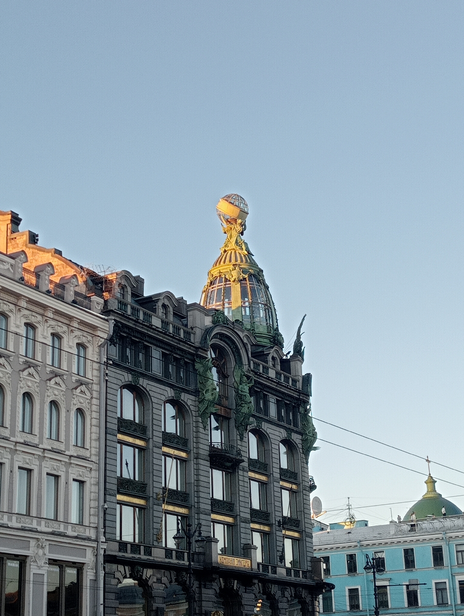 The image shows the Singer House, also known as the Dom Knigi (House of Books), located on Nevsky Prospekt in Saint Petersburg, Russia. The building features a distinctive glass dome topped with a sculpture of an eagle and a globe. The architecture is ornate, with classical and art nouveau elements. Sunlight illuminates the upper portions of the building, casting a warm glow. In the foreground, the facade of the Singer House is visible, with its numerous windows and decorative details. To the right, other buildings, including a blue one with a green dome, are partially visible. The sky is clear and pale blue, indicating daylight. There are no people visible in the image.