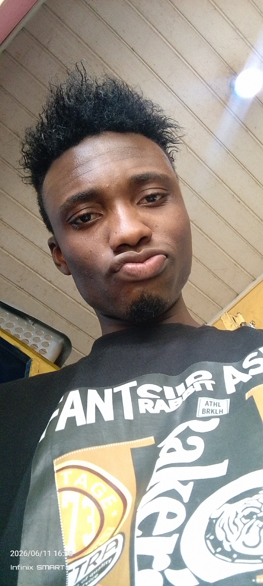 A medium shot photo is captured from a low angle, focusing on the face and upper body of a young Black man. He is looking directly at the camera with a pout, his lips pursed and his eyes slightly narrowed. His hair is styled in a short, spiky afro. He has a goatee. He is wearing a black t-shirt with white lettering and graphics. The background consists of a textured, light-colored ceiling with visible slats. To the left of the man, a portion of what appears to be a metal grate or ventilation system is visible, colored yellow and dark blue. The lighting suggests an indoor environment, possibly a public space or a casual setting. The timestamp "2026/06/11 16:08" and "Infinix SMART 3" are superimposed in the lower left corner of the image.