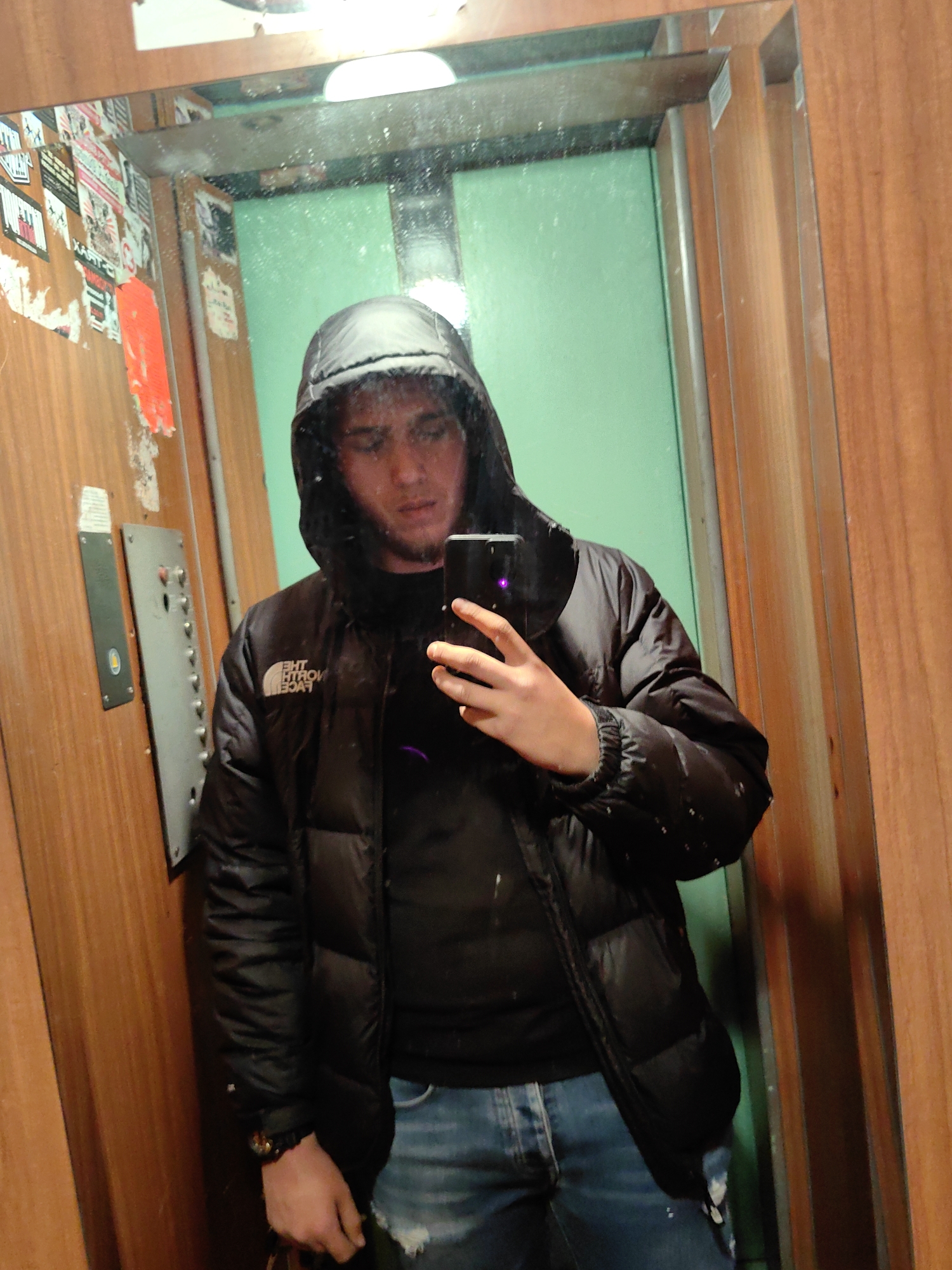 A man wearing a black hooded jacket and blue jeans is taking a selfie in a mirror. The mirror is in a lift/elevator, judging by the elevator panel and the stickers on the wall. The elevator has wood paneled sides and a green back wall. The scene could be in Sofia, Bulgaria, but there is no concrete evidence for that based on the image alone.