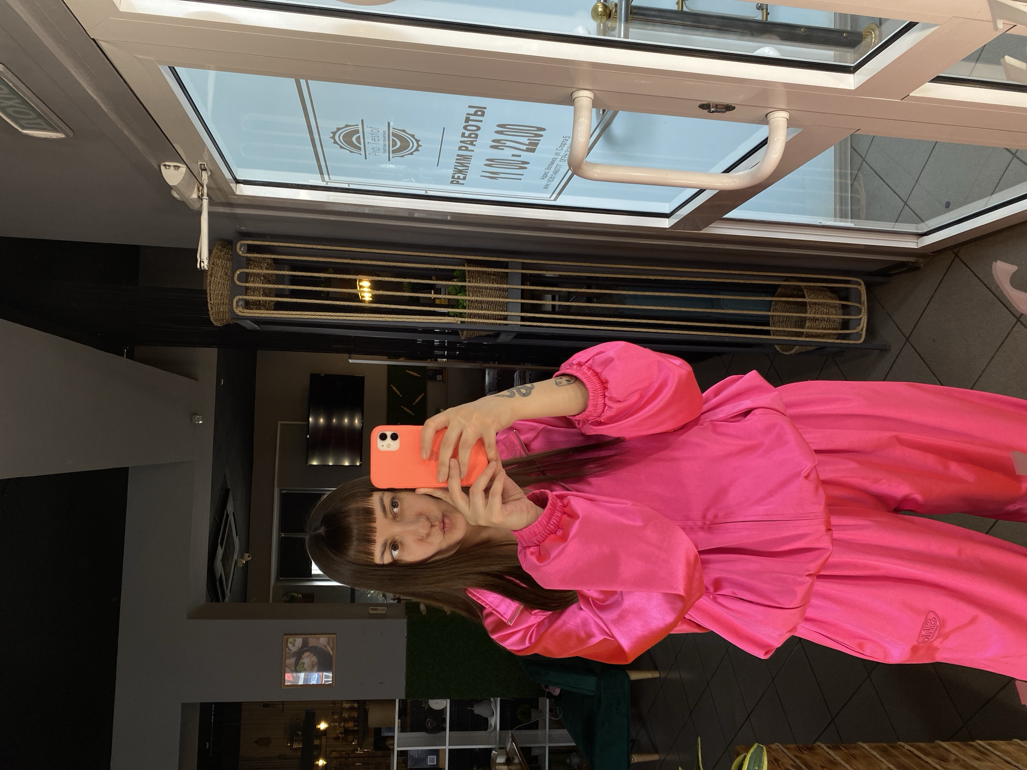 A person in a pink outfit is taking a selfie in front of a glass door. The door has a sign indicating "Pro Testof" and business hours from 11:00 to 22:00. The sign also includes the address: Votkinsk, Sports Street, 5, with tax identification numbers. The interior of the building is visible through the door.