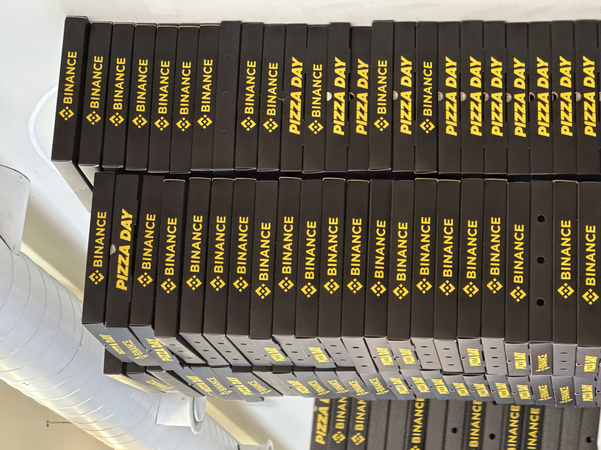 The image shows a collection of black pizza boxes stacked on a shelf. The boxes are printed with the "Binance" logo and the phrase "Pizza Day" in yellow lettering. The boxes appear to be arranged in two tiers, with some boxes partially obscuring others. The image indicates that the event is sponsored by Binance and celebrates "Pizza Day". The location is not specified in the picture, and there is no indication that the image was taken in Yerevan, Armenia.