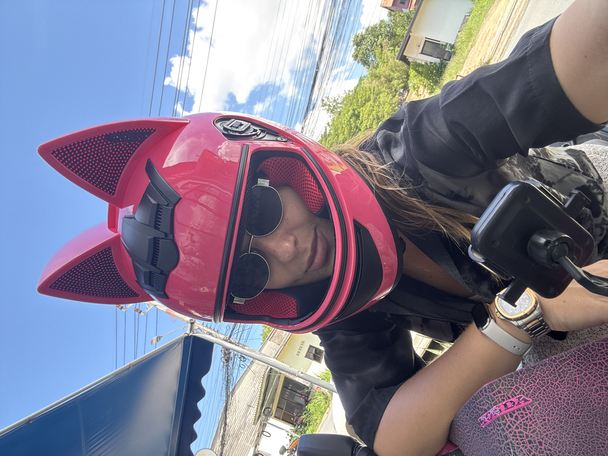 A woman wearing a pink motorcycle helmet with black cat ears is captured in a selfie. She has on sunglasses and a black top. Her left arm rests on a pink object with a textured pattern and a prominent "HONDA" logo in pink. She is wearing a silver watch on her left wrist. The background shows a street scene with buildings, trees, power lines, and a blue awning, suggesting an outdoor, possibly urban or suburban, environment in Thailand, specifically in the area of Ban Bang Rak. The sky is bright blue with some scattered clouds.