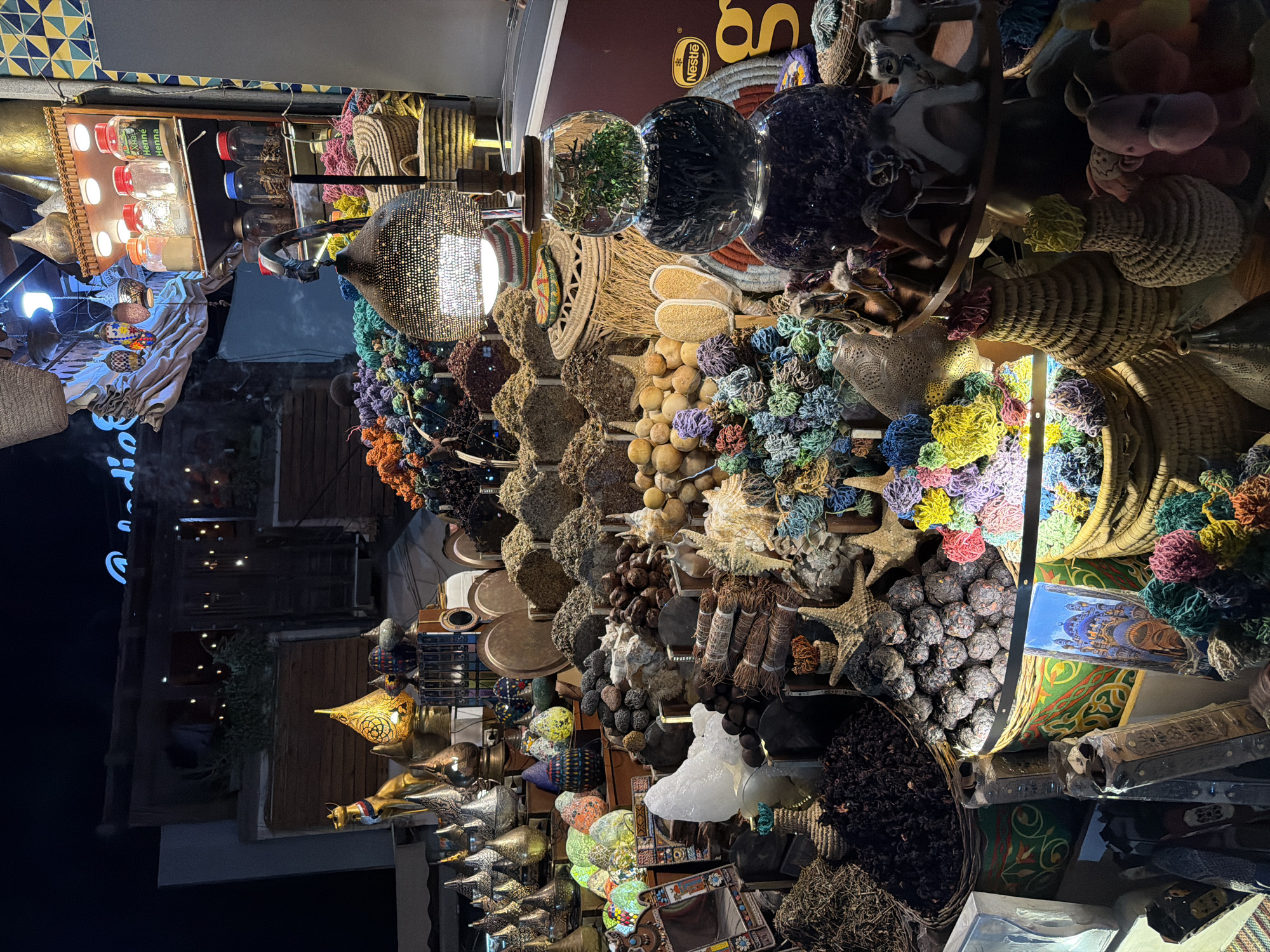 The image depicts a nighttime scene in Sharm El Sheikh, Egypt, likely at a market or shop selling spices and artisanal goods. The foreground is dominated by a vibrant display of various items. Baskets overflowing with colorful, intricately arranged spices or dried flowers in shades of blue, purple, green, yellow, orange, and red are prominently featured. Alongside these, piles of dried herbs, what appear to be date fruits, small round objects that could be dried fruits or seeds, and larger, smooth stones are visible.
A decorative lamp with a perforated metal shade hangs above the display, casting patterned light. To the right, a stack of three clear glass spheres contains dried herbs and what appears to be dark, finely cut plant material. A Nestle logo is visible on a dark red background on the right side of the frame.
In the mid-ground, various decorative objects are displayed, including metallic lamps in conical and rounded shapes, some adorned with intricate patterns. Colorful, textured glass lamps in egg shapes are also present, along with intricately carved mirrors. Further back, shelves are stocked with jars containing powders and dried plant materials, labeled with what appears to be "Henna."
The background shows the facade of a building with illuminated windows and decorative lighting, suggesting an establishment open at night. A neon sign, partially obscured, displays blue lettering that might spell out a name. The overall impression is one of a bustling, atmospheric marketplace offering a wide array of sensory experiences. There are no clearly identifiable people engaged in specific actions.