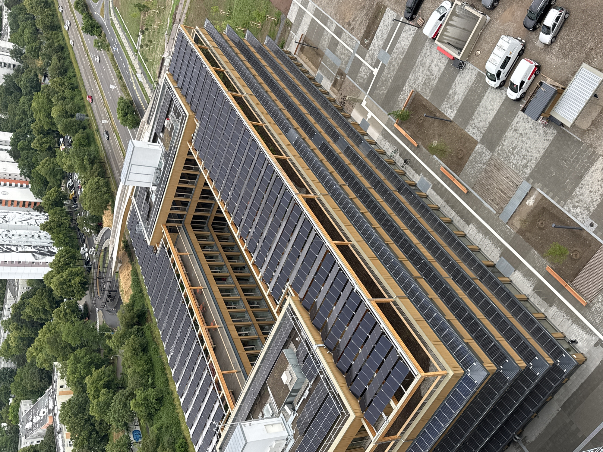 This aerial image, taken in Mannheim, Germany, captures a modern, multi-story building with extensive solar panel installations on its roof and facade. The building features a U-shaped design with a courtyard, characterized by its wooden exterior and numerous windows. Rows of solar panels are visible on multiple levels, covering large sections of the roof and extending down the sides of the building.
The surrounding environment includes a highway with several cars driving on it, green spaces with trees, and a paved area in front of the building. Several vehicles, including white vans and cars, are parked on the paved area. The overall scene suggests a contemporary urban setting with a focus on sustainable energy infrastructure.