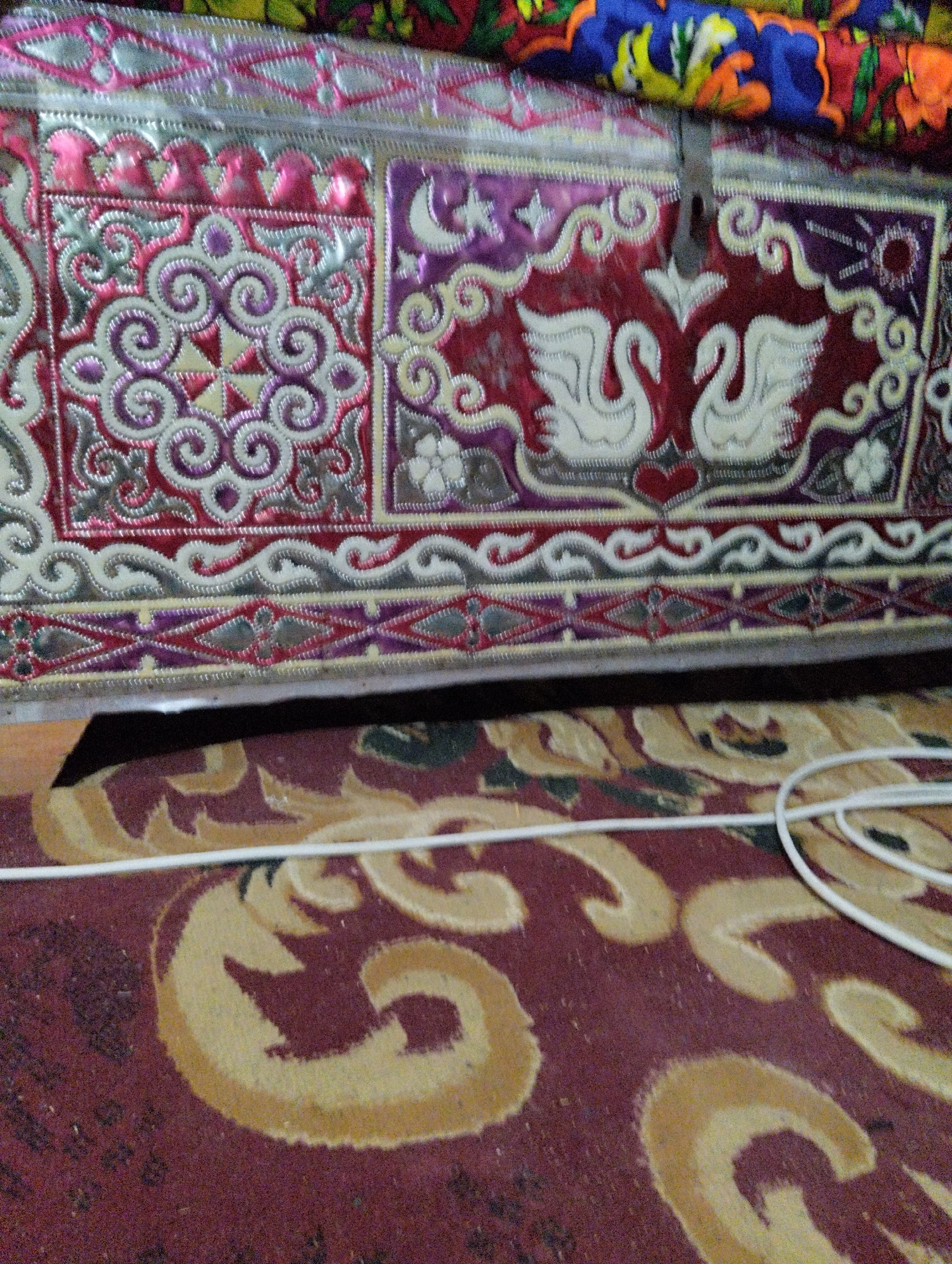 The image shows a close-up of a decorative wooden chest with ornate metalwork, situated on a patterned rug. The chest features intricate designs in red, silver, and purple, including floral motifs and two white swans facing each other. The rug is a deep red with large, swirling yellow patterns. A white cable is visible in the foreground, partially covering the rug. The scene is indoors, with the top of the chest revealing colorful fabric. The location is identified as Qanlikul, Uzbekistan. There are no people or animals visible in the frame, and no actions are being performed. The overall impression is one of a decorative interior scene, likely a room within a home or a traditional dwelling.