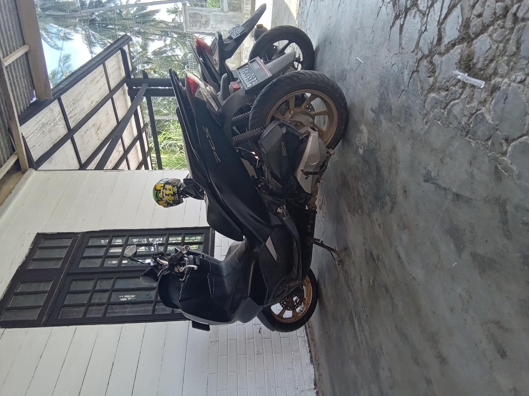 The image depicts an outdoor scene in Lhokseumawe, Indonesia, beneath a covered structure. Two motorcycles are parked on a grey concrete surface, adjacent to a white building.
The prominent motorcycle in the foreground is a black Honda PCX scooter, identifiable by the "PCX" emblem on its side and its gold-colored alloy wheels. It is resting on a side stand. A yellow and green helmet, marked with "GM" and "46", is placed on the rear seat of this scooter. The visible Indonesian license plate reads "BL 6313MA". Partially obscured behind it is a second, similar black scooter, with its rear wheel, taillight, and a partially visible license plate (starting with "BL") discernible.
To the left, a white building with horizontal paneling or painted brickwork features a tall, dark-framed window composed of multiple small panes. Overhead, a covered structure with a corrugated metal roof is supported by dark beams and posts; its underside reveals white panels.
The concrete ground exhibits various stains, cracks, and textured patterns, particularly on the right side where a section appears to be stamped or patterned. Small pieces of white plastic debris are visible on the ground.
In the background, beyond the covered area, tall palm trees and other green foliage are visible against a bright blue sky with scattered white clouds. A concrete wall or fence is also present in the distant background to the right. No individuals are present in the frame.