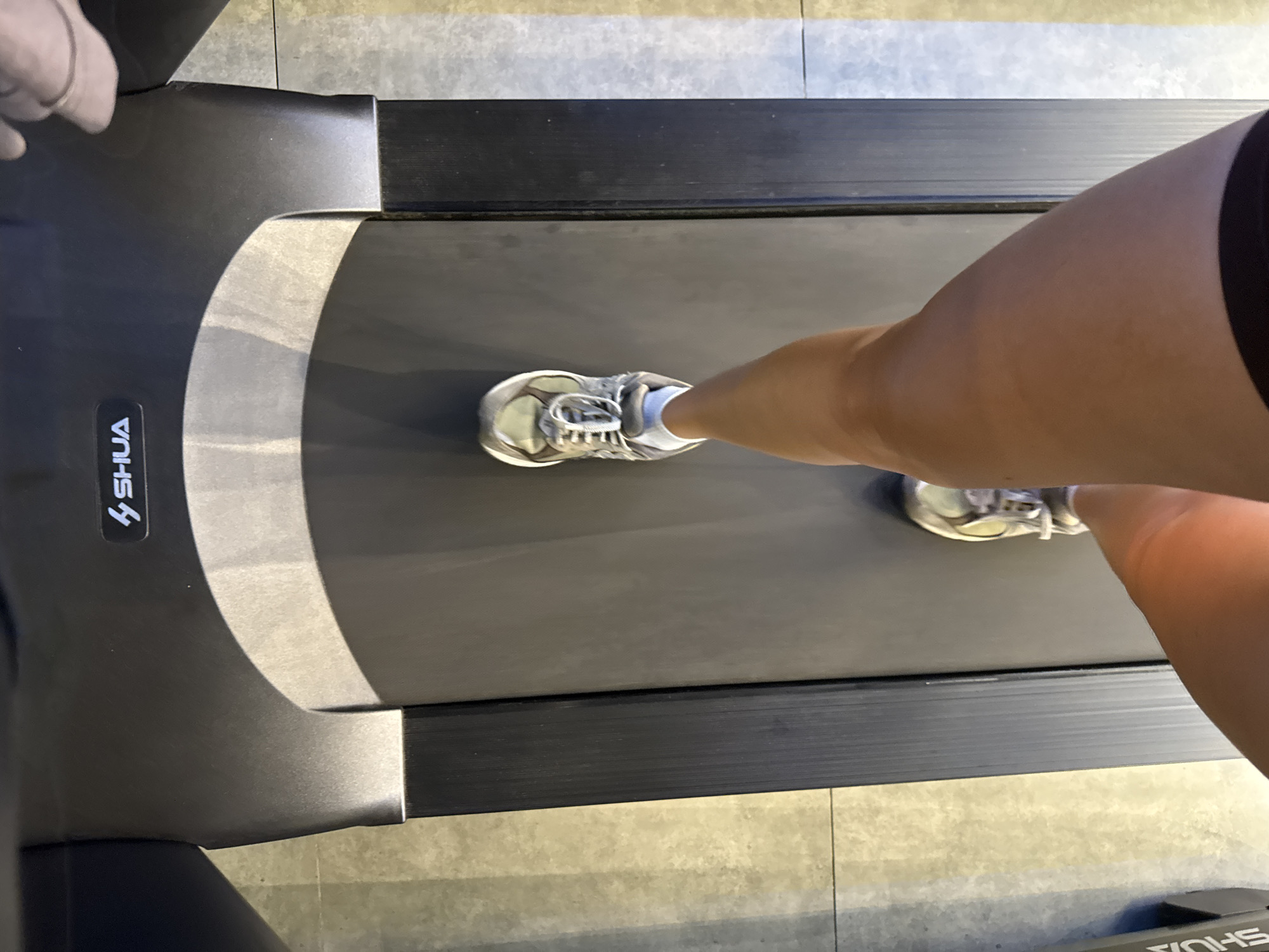 In Nam Nha Trang Ward, Vietnam, a person's legs are shown from a first-person perspective as they walk on a treadmill. The treadmill is black with a silver accent and features the "SHUA" logo. The person is wearing grey athletic shoes with white laces and light blue socks. The floor around the treadmill is tiled in a beige and grey pattern. The lighting suggests an indoor setting, possibly a gym.