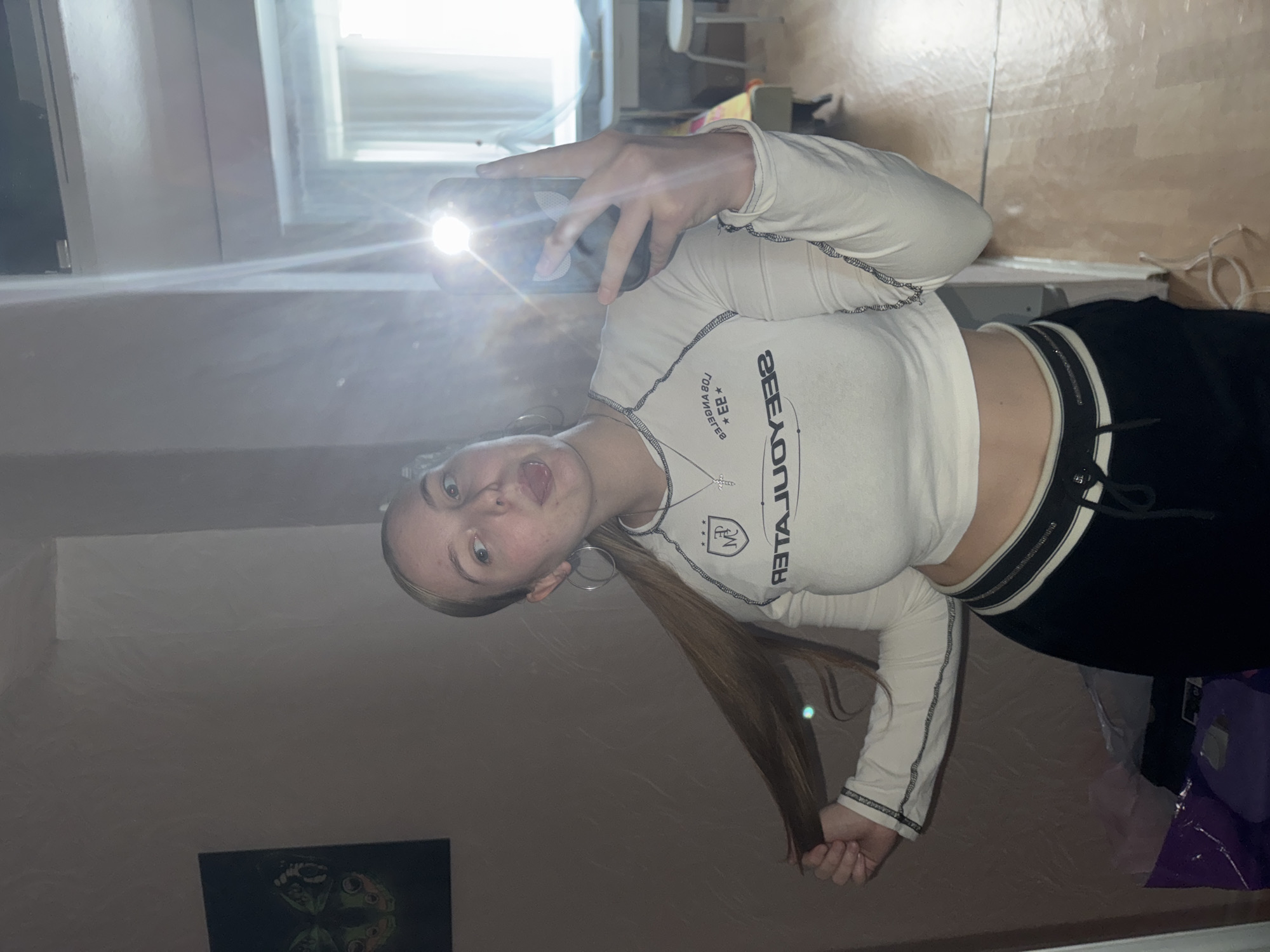 The media file shows a young person taking a selfie in a room. The person is wearing a long-sleeved white crop top with black stitching and black pants with a white stripe and zipper detail. They have long brown hair and are wearing large hoop earrings and a silver necklace with a cross pendant. The person is making a playful expression, sticking their tongue out. A phone is held in their hand, with its flash illuminating the scene.
In the background, a large mirror reflects the room, revealing a window and parts of furniture. The reflection also shows a framed picture on the wall, featuring a design that appears to be a skull or a similar graphic. The overall lighting suggests it is daytime, as indicated by the bright light from the window. The setting seems to be a personal living space, likely a bedroom.
Visible text on the person's shirt appears to be "SEE YOU LATER" and "LOS ANGELES". The image is oriented sideways. The location context provided is Perm, Russia.