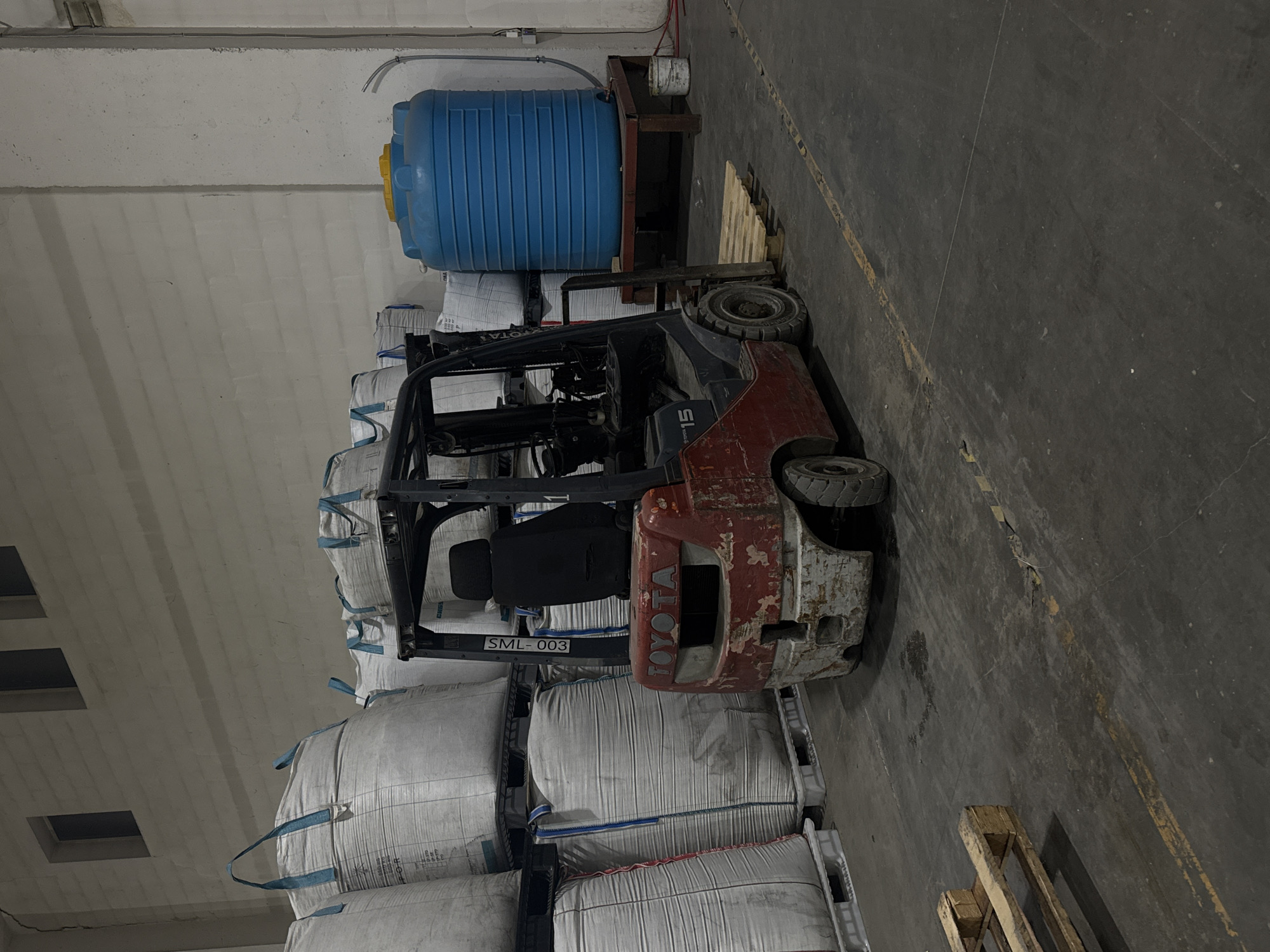 The image depicts an indoor warehouse setting, possibly located in Perm, Russia. A red Toyota forklift truck, showing signs of wear and rust, is positioned amongst stacks of large white bags. The bags are arranged on pallets. A blue water tank is situated near the forklift, elevated on a platform. The warehouse appears to be well-lit with grey flooring. The forklift has "SML-003" printed on the side.