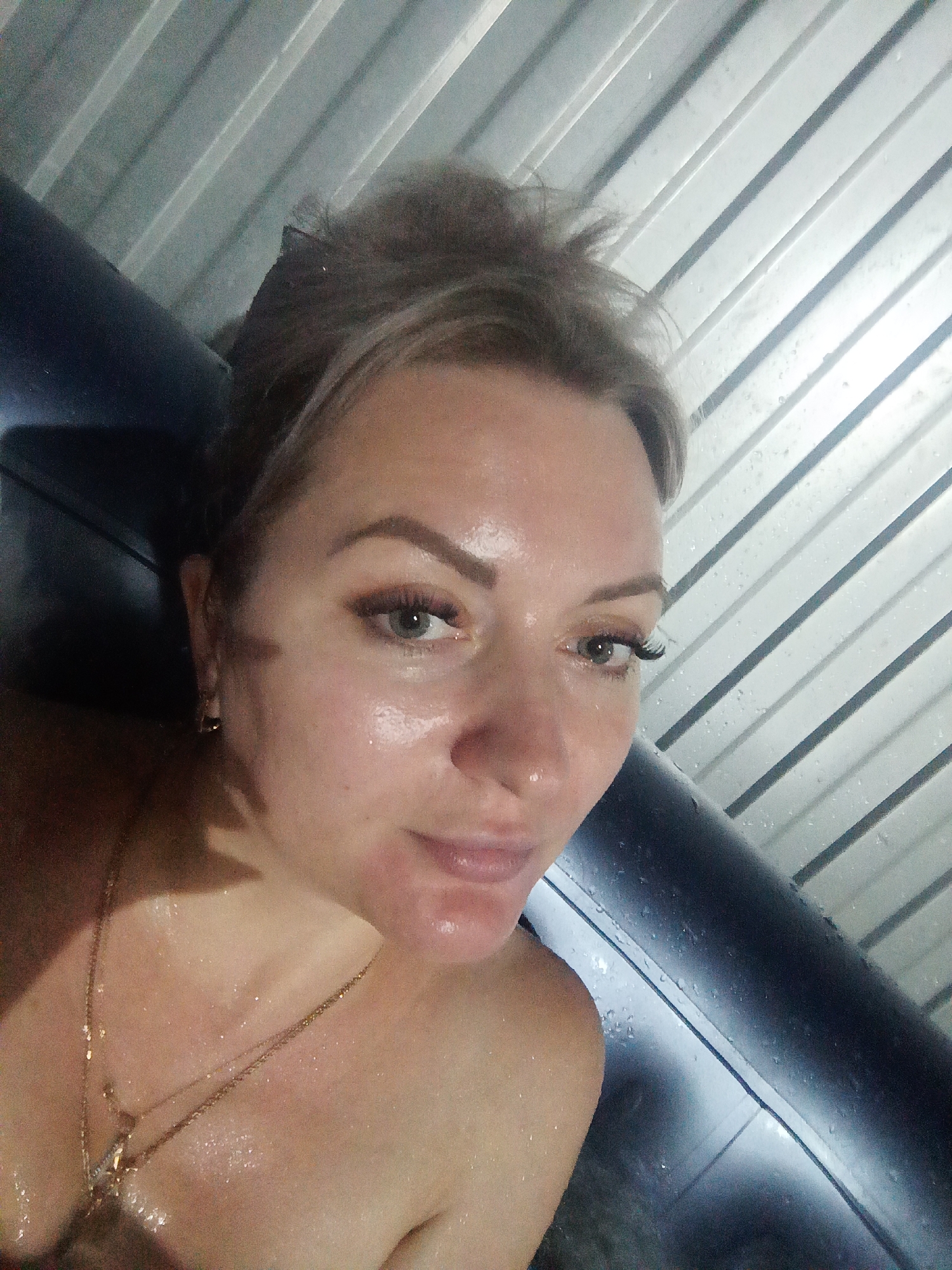 The image is a selfie taken by a woman in what appears to be a steam room or sauna. The woman has blonde hair pulled back, light-colored eyes, and is wearing makeup, including false eyelashes. She is wearing a gold necklace. Her skin appears to be glistening with sweat or water. The background consists of a ribbed metal ceiling and a dark-colored, curved surface, possibly a bench or wall. There are water droplets visible on the ceiling. The overall impression is that the woman is in a humid, enclosed environment, likely a spa or a similar facility. The location is identified as the city of Sorochinsk, Russia.
