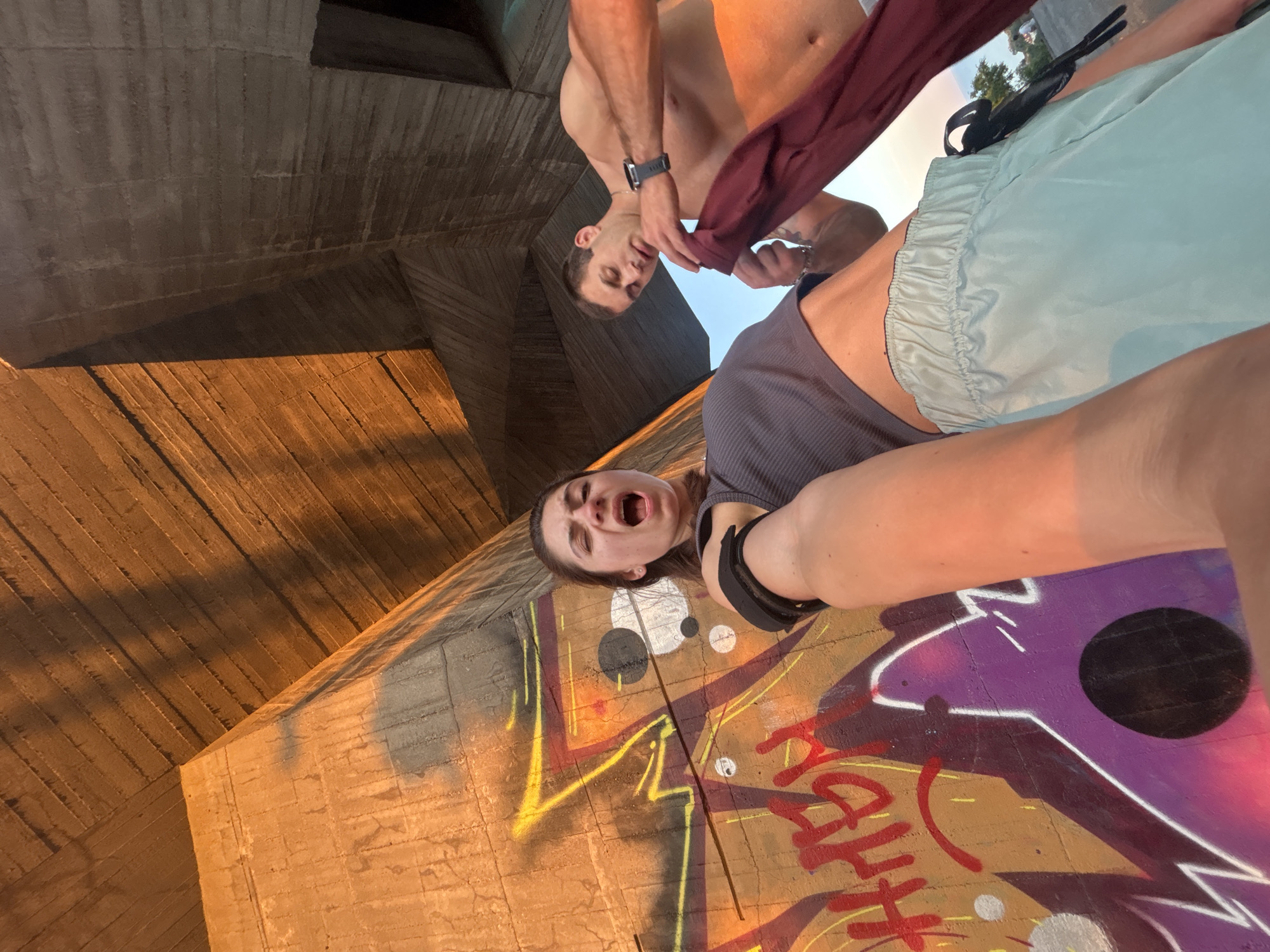 The image depicts a low-angle, wide shot of two individuals standing in front of a graffiti-covered concrete structure, likely under a bridge, possibly in Varna, Bulgaria. The setting appears to be outdoors, with the sun casting warm, orange light on the concrete and creating strong shadows.
In the foreground, a woman is captured from a low angle, with her face contorted in a yell or scream, eyes partially closed, and mouth wide open. She is wearing a dark grey, ribbed tank top and light blue athletic shorts. Her right arm is extended towards the camera, holding the device capturing the image. A black strap is visible on her upper right arm.
Behind and to the right of the woman, a shirtless man is looking down as he pulls a dark red garment, possibly a t-shirt, over his head. He is wearing a dark watch on his left wrist.
The concrete structure behind them is heavily graffitied with vibrant colors, including red, yellow, white, and purple. The graffiti appears abstract and stylized. The underside of the concrete structure is angled and textured, suggesting it is part of a large man-made construction. The overall impression is one of urban exploration or a candid moment captured in a somewhat gritty, artistic environment.