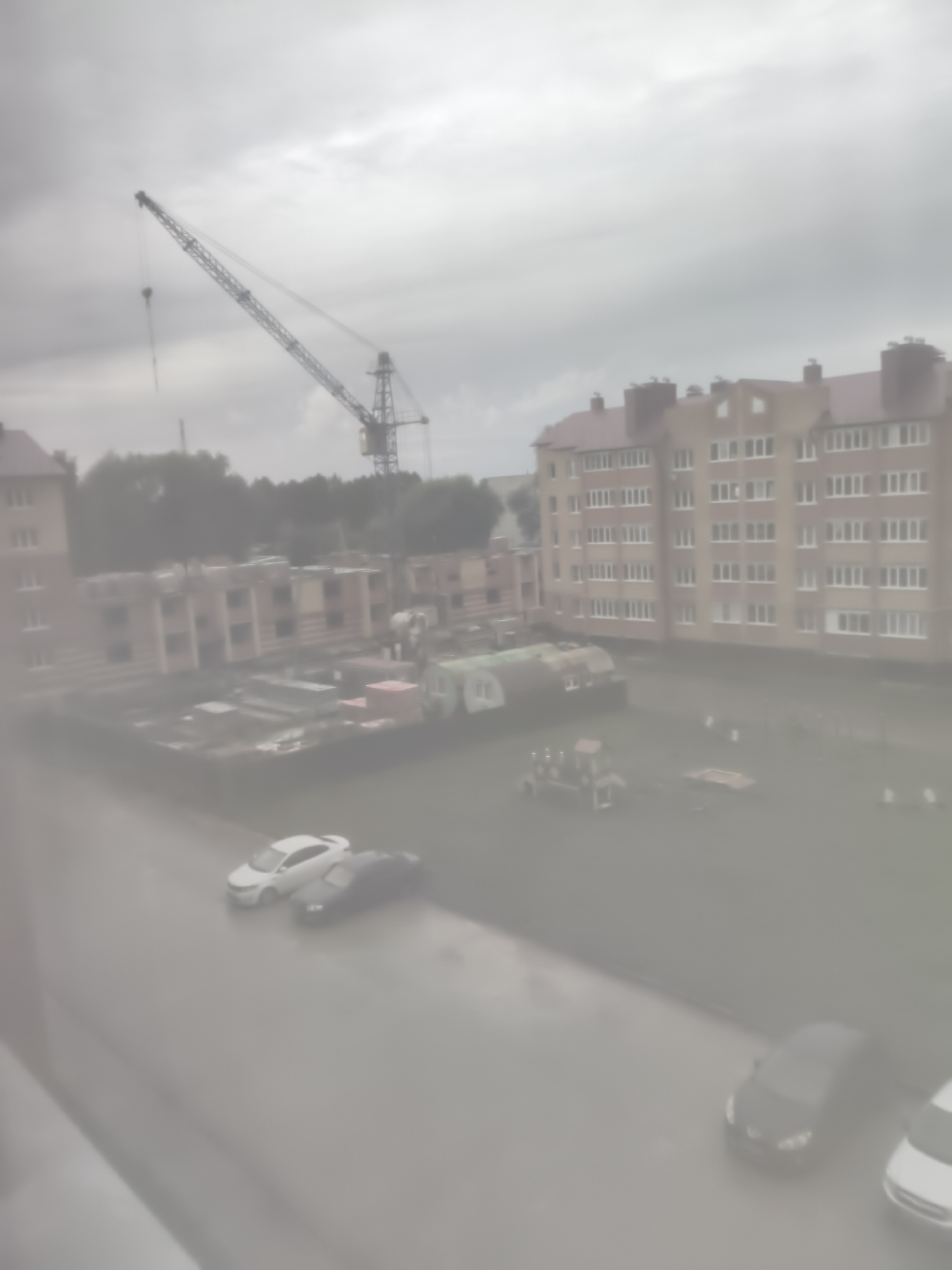 The image shows a construction site with a crane in the background, a residential building, and a street with parked cars. The sky is overcast, and the scene appears to be in an urban or suburban area. The buildings appear to be multi-story residential structures, possibly apartment complexes. There is also a playground visible in the courtyard. The cars parked on the street are a white sedan and a dark-colored sedan. The overall impression is of a typical day at a construction site in a residential neighborhood, with the weather being somewhat dull. It is difficult to ascertain the exact location within Tsivilsk, Russia, without further context.