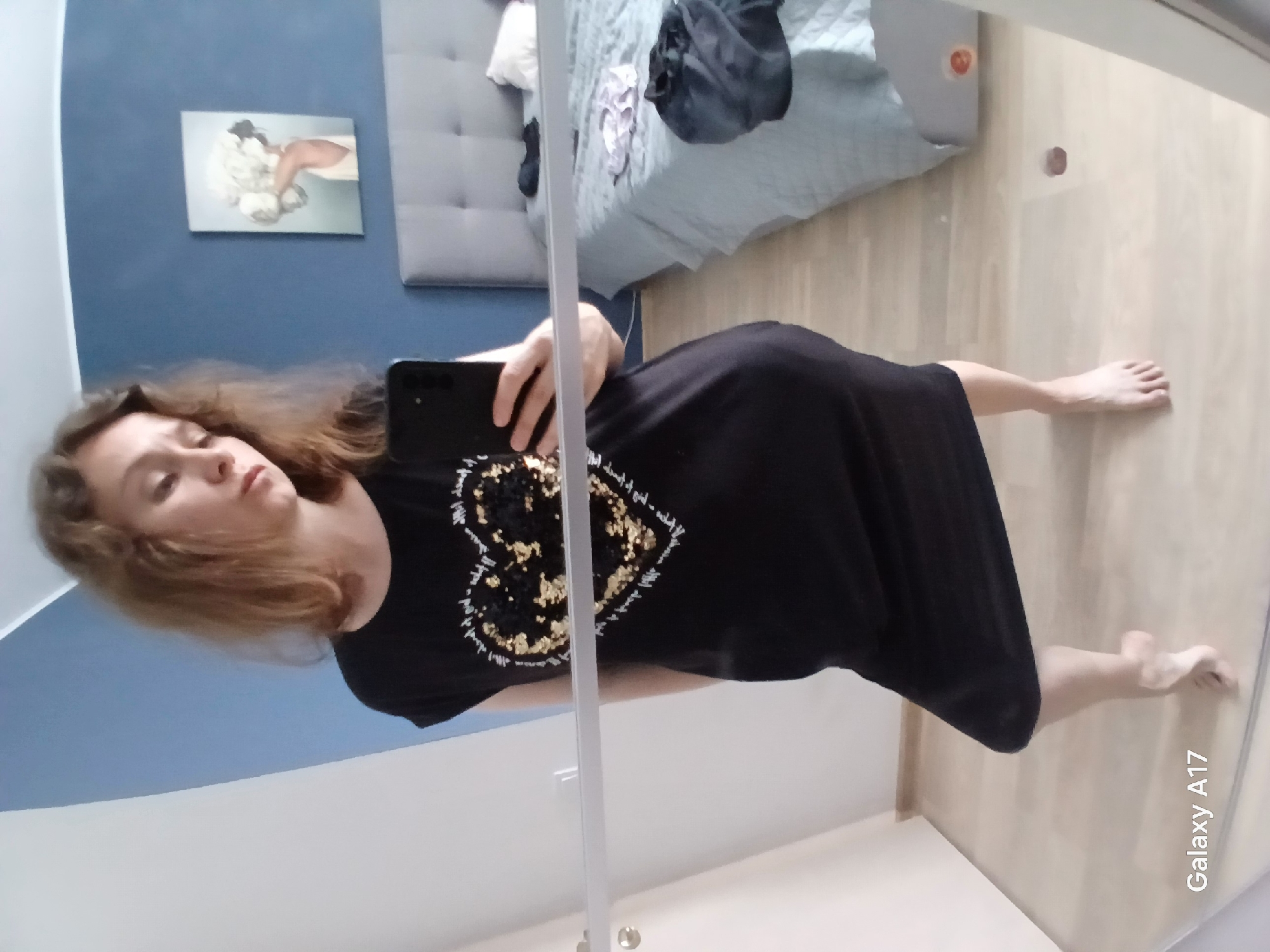 A woman is taking a selfie in what appears to be a bedroom. She is wearing a black t-shirt with a gold heart design. She is standing in front of a mirror. The background shows a bed with a blue comforter, a painting on the wall, and light-colored wooden floors. The phone being used to take the photo displays the text "Galaxy A17". The city of Yekaterinburg, Russia, cannot be determined from the image.