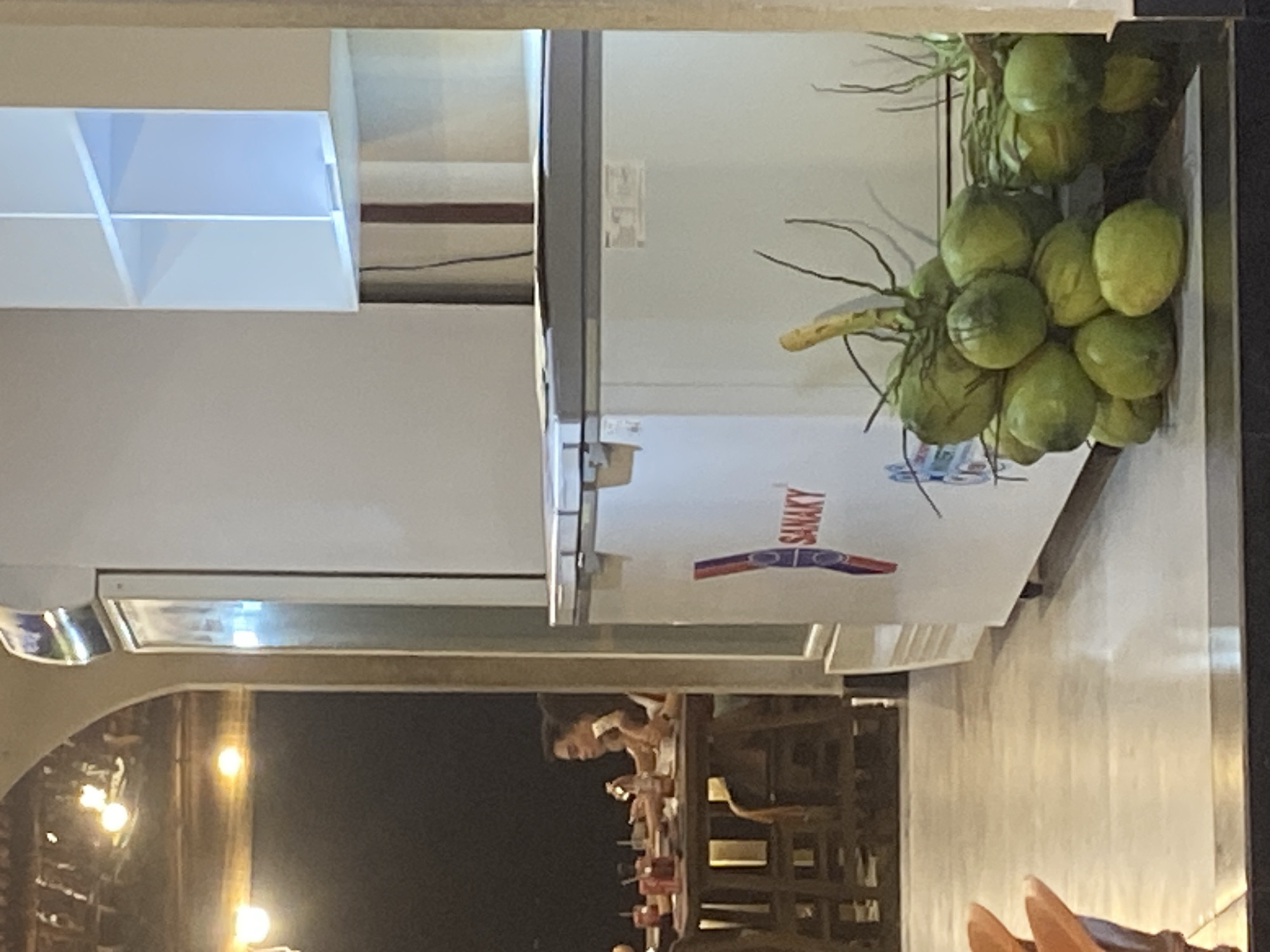In Mũi Né, Vietnam, a food establishment displays a chest freezer adorned with the "SANAKY" logo. A cluster of green coconuts rests on the edge of the freezer, with some branches extending upwards and to the right. In the background, two individuals are seated at a wooden table, appearing engaged in conversation or dining. The setting features overhead lighting and an arched ceiling structure.