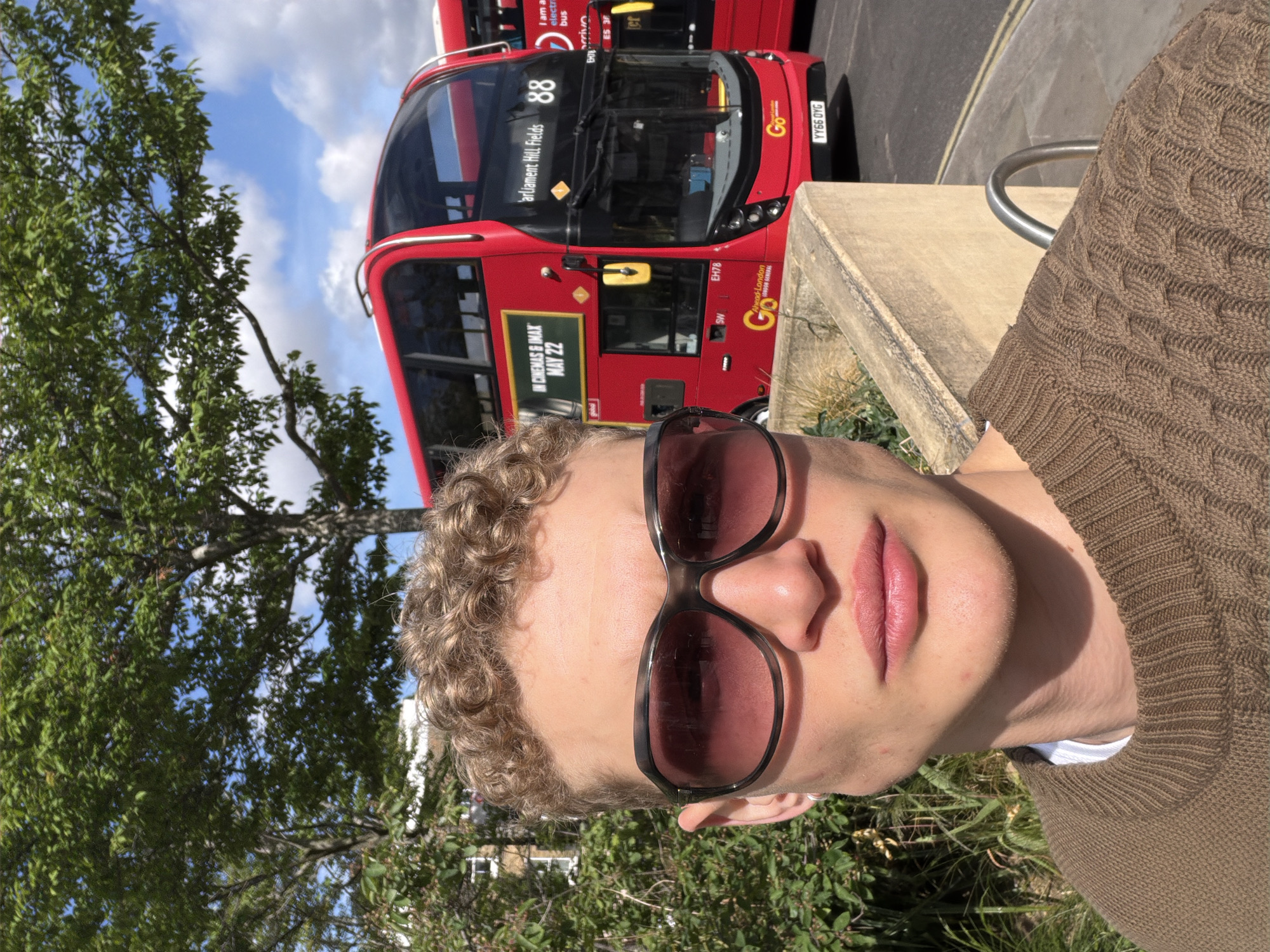 The image is a close-up selfie of a light-skinned person with short, curly blonde hair and wearing sunglasses, standing outdoors on a sunny day. Behind them is a red, double-decker Go-Ahead London bus with the number "88" and the destination "Parliament Hill Fields" displayed on its front. The bus is identified as an electric vehicle. The side of the bus has an advertisement for a movie showing in cinemas and IMAX, with a date of May 22. A tree with green leaves is partially visible to the left. Street furniture, including what looks like a concrete bench and metal railing, is also present. The license plate of the bus is YY66 DYG.