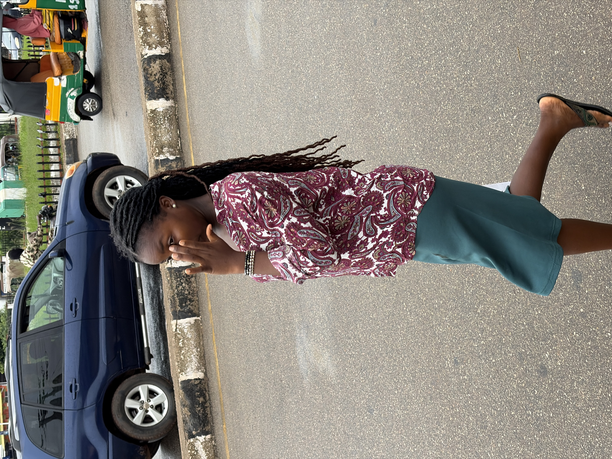 A young Black woman stands outdoors on a paved surface, possibly a road or sidewalk, with a dark blue SUV to her left and a yellow and green auto-rickshaw in the background. She is wearing a patterned long-sleeved blouse and a solid dark green skirt, and black flip-flops. Her hair is styled in long dreadlocks. The woman has her right hand raised to her face, obscuring her mouth and nose, with her fingers splayed. Her left arm is bent at the elbow, with a beaded bracelet on her wrist. The overall impression is one of a candid moment, perhaps a pose for a photograph, with the woman appearing to look towards the camera. The lighting suggests it is daytime, and the weather appears to be clear. The presence of the vehicles and the paved area indicate an urban or semi-urban setting. The auto-rickshaw is a common mode of transport in Nigeria.