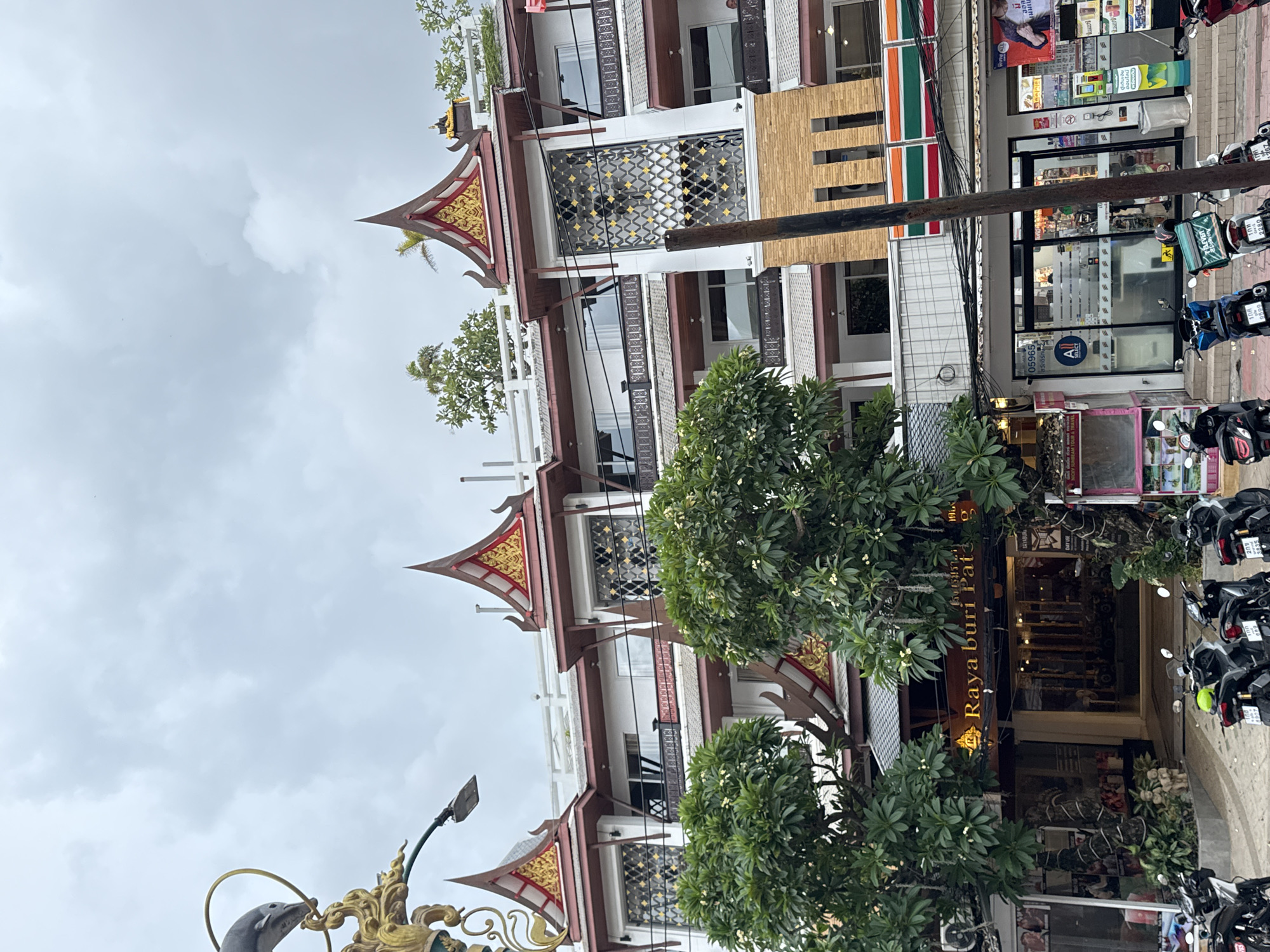 A building with traditional Thai-style tiered roofs, decorated with gold and red accents, stands under an overcast sky in Patong, Thailand. The building features white walls and multiple levels with balconies and dark-colored railings. Trees with green foliage and white flowers partially obscure the lower floors. Several parked motorcycles are visible in front of the building, which appears to be a hotel or resort, indicated by a sign reading "Raya Buri Patong". Adjacent to the hotel entrance, a 7-Eleven convenience store is visible with its distinctive red, green, and orange signage. The street is paved, and electrical wires are strung across the scene.