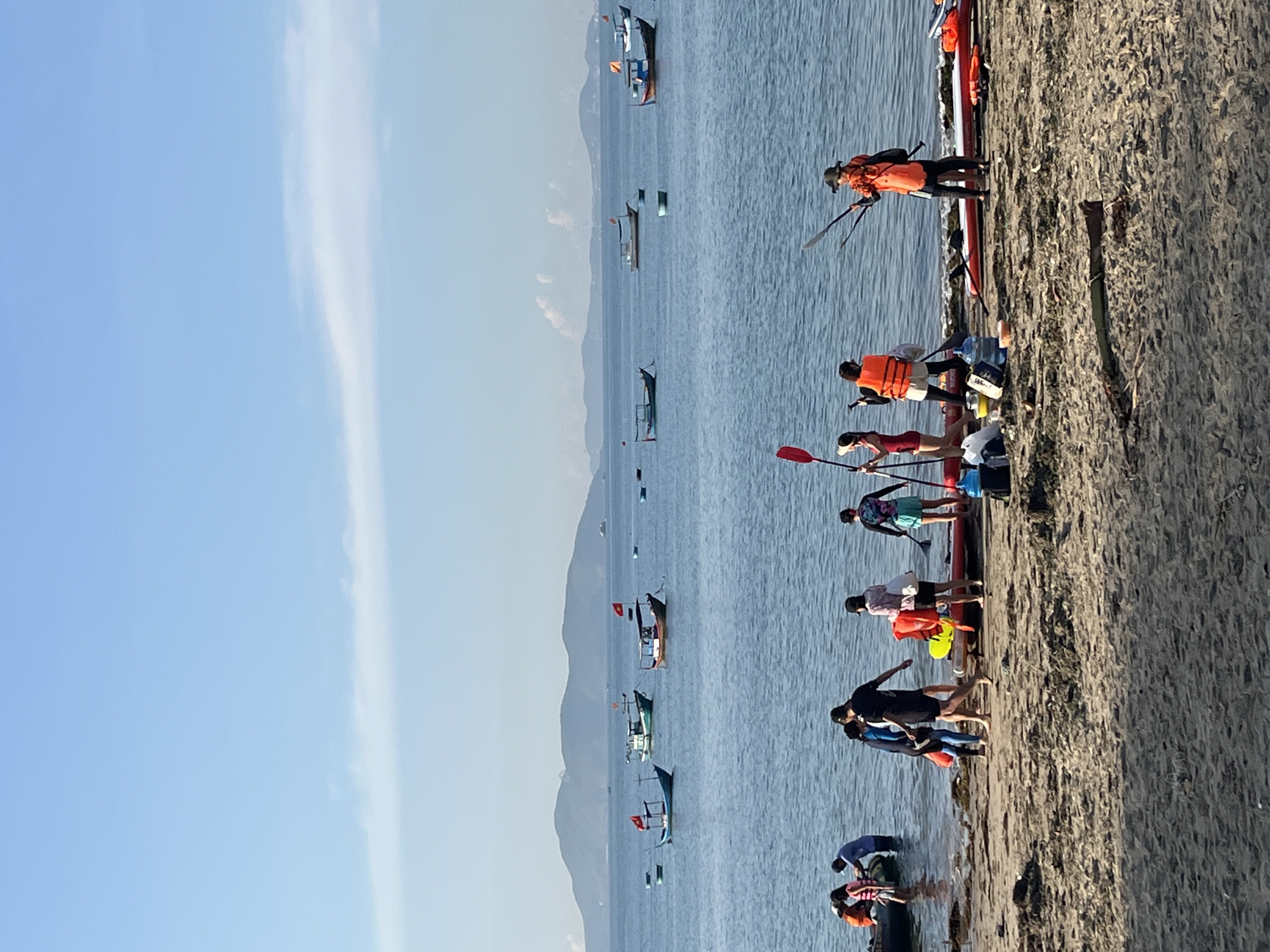 The image depicts a coastal scene in Bac Nha Trang Ward, Vietnam. The horizon is dominated by a light blue sky, with a tall, thin cloud formation extending vertically. Below the sky, the sea is visible, with small islands or mountains in the distance. Several boats are scattered across the water.
Along the shoreline, a group of people are gathered. They appear to be involved in watersports activities, potentially kayaking or paddleboarding, as evidenced by the presence of paddles and a long, narrow watercraft on the shore. Some individuals are wearing life jackets. The ground near the water's edge is a sandy texture.