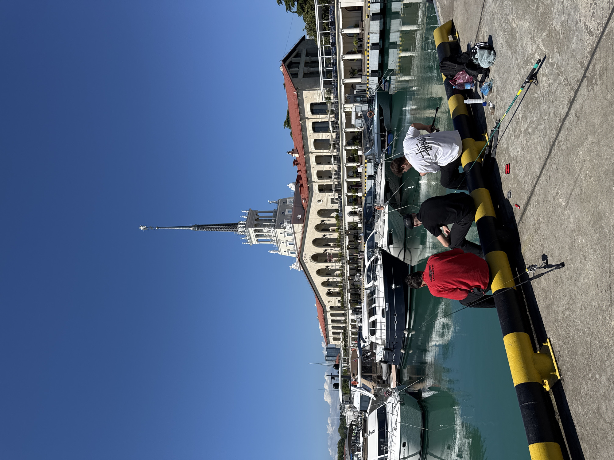 The image, taken on a clear and sunny day, depicts a scene at what is identifiable as the Sochi Sea Port in Russia. A wide, bright blue sky dominates the upper portion of the frame, overlooking calm, turquoise-green water.
On a concrete pier in the foreground, three men are observed fishing. They are positioned with their backs to the camera, leaning over the edge of the pier. One individual wears a white t-shirt with a black "STÜSSY" logo on the back, another is in a black long-sleeved shirt, and the third wears a red long-sleeved shirt featuring an "UMBRO" logo. Fishing rods are visible, with one propped against a black and yellow striped bollard, and another being actively held. Adjacent to the person in white, a black backpack, a grey cloth, and several plastic bottles are placed on the pier.
The background features a grand, ornate building with classical architectural elements, including a series of arches, balconies, and a distinctive red tiled roof. Behind this structure, a prominent white spired tower, topped with a star, rises into the sky, a notable landmark of the Sochi Sea Port Terminal. Numerous yachts and boats, varying in color from white to dark-hulled, are docked in the marina, their forms and the surrounding buildings reflecting clearly in the still water. Text "P-04-0994" and "Сочи" (Sochi in Cyrillic) are partially visible on the hull of a black boat. The bright, direct sunlight suggests it is midday or early afternoon, contributing to a serene and leisurely atmosphere.