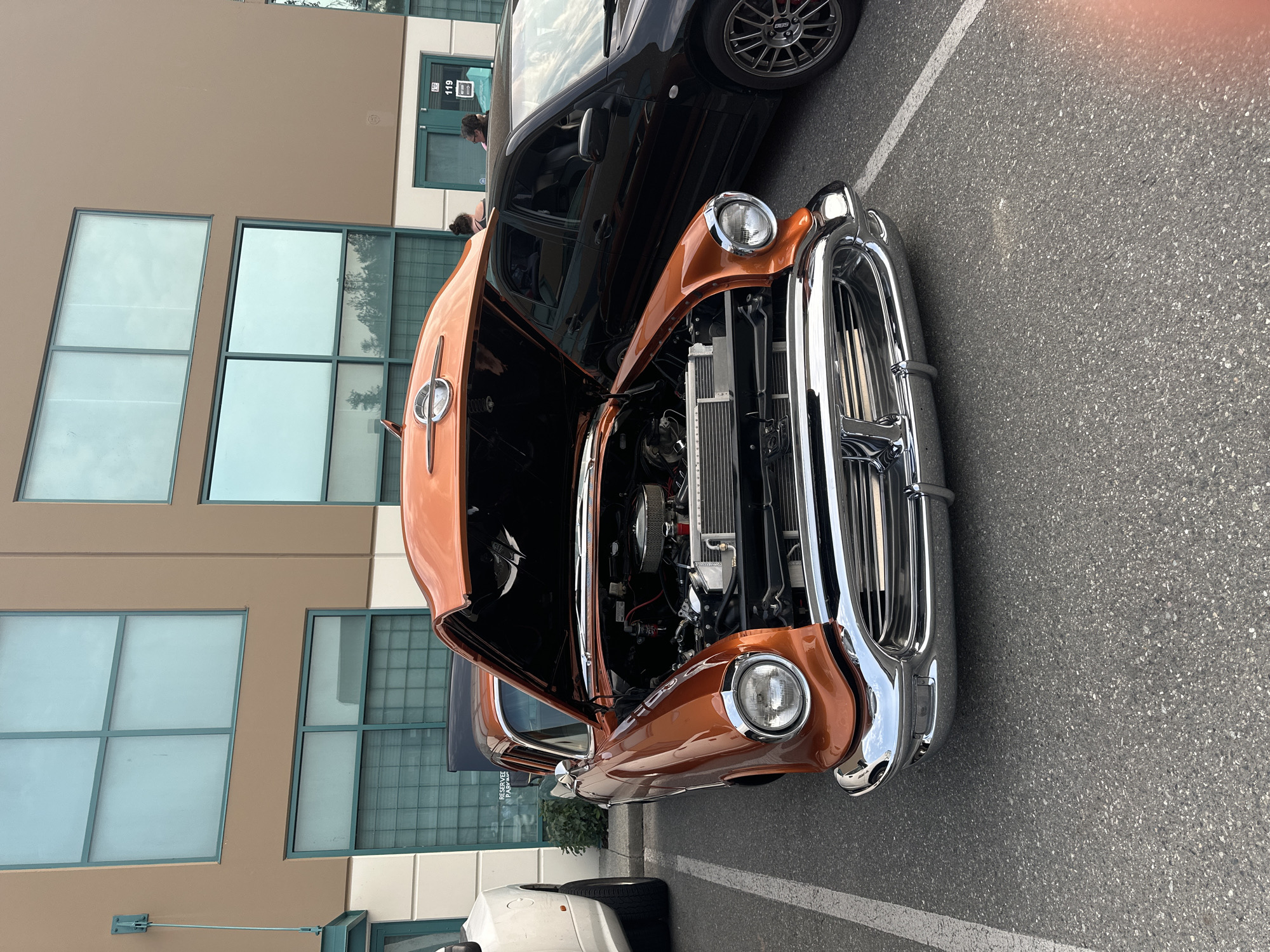 An orange classic car with its hood open is parked in a lot, possibly at a car show or event. The car features chrome accents, including the grille and headlights. Behind the classic car, a black sedan is parked, and a portion of a white vehicle is visible to the left. The background consists of a modern building with large, rectangular windows and tan siding. Two individuals are visible standing near the black sedan and the building entrance. The ground is paved asphalt with white parking lines. The scene appears to be outdoors, under daylight conditions.