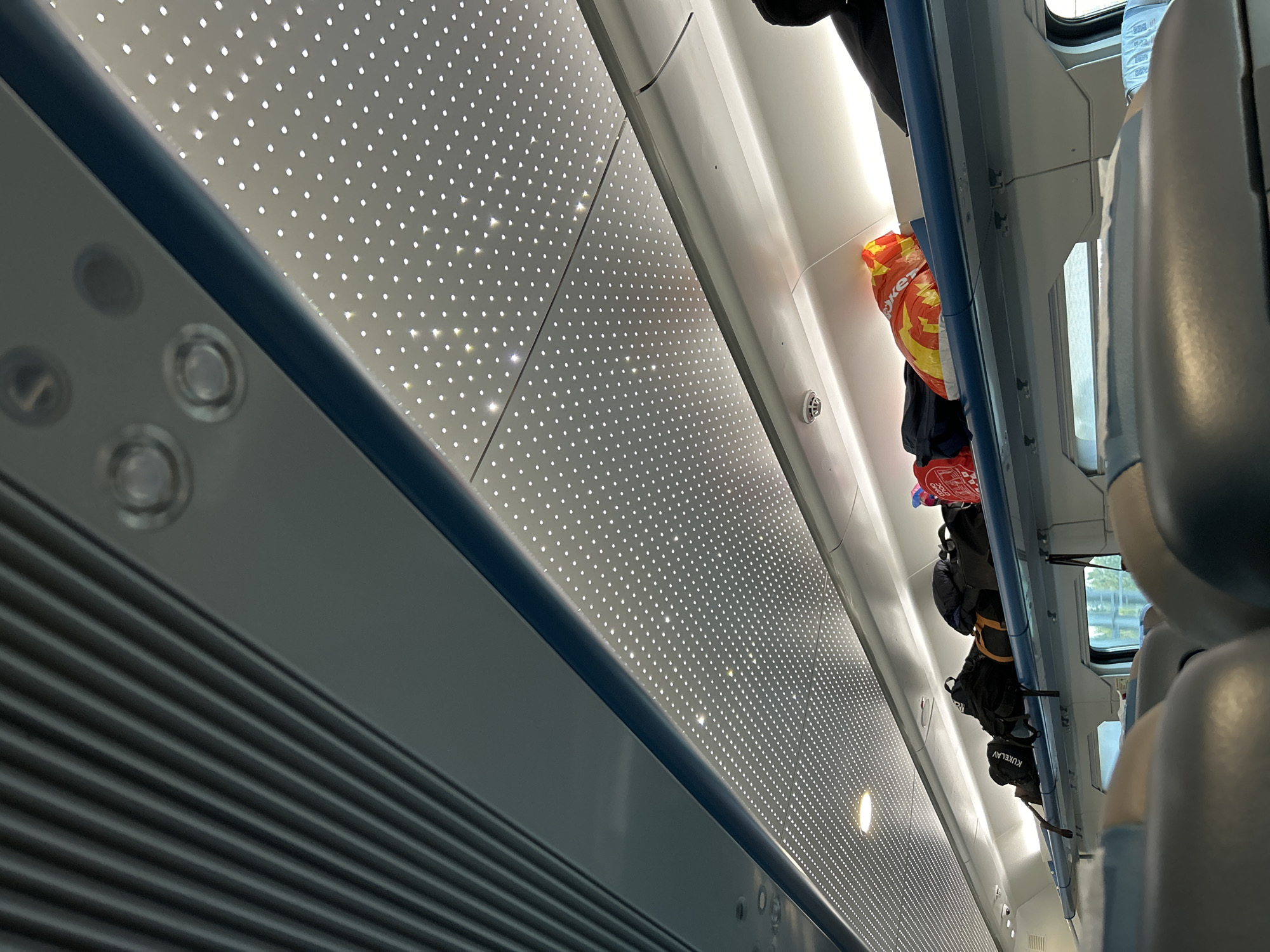 The image displays the interior of a modern train carriage, captured from a low, tilted angle looking across the aisle. On the left side, a blue trim outlines a panel featuring three circular fixtures, positioned above a gray panel with horizontal grilles, likely an air conditioning vent.
The prominent ceiling and upper wall sections are white and perforated, embedded with numerous small, bright LED lights arranged in a grid pattern, illuminating the space.
On the right, an overhead luggage rack is visible, holding several bags and backpacks. A black backpack clearly shows the white text "KUKELAV." Another item is an orange and yellow plastic bag with partially visible Cyrillic text, which appears to be "Жет," likely part of "Маркет" (meaning 'Market'). A clear plastic water bottle is also stowed on the rack. Through a window partially seen on the far right, a blurry green outdoor landscape suggests the train is in motion during daylight. Parts of light-colored seats are visible in the foreground on the right.
The scene depicts typical travel activity within a contemporary train setting. The visible Cyrillic text suggests the location might be in a Slavic-speaking region.