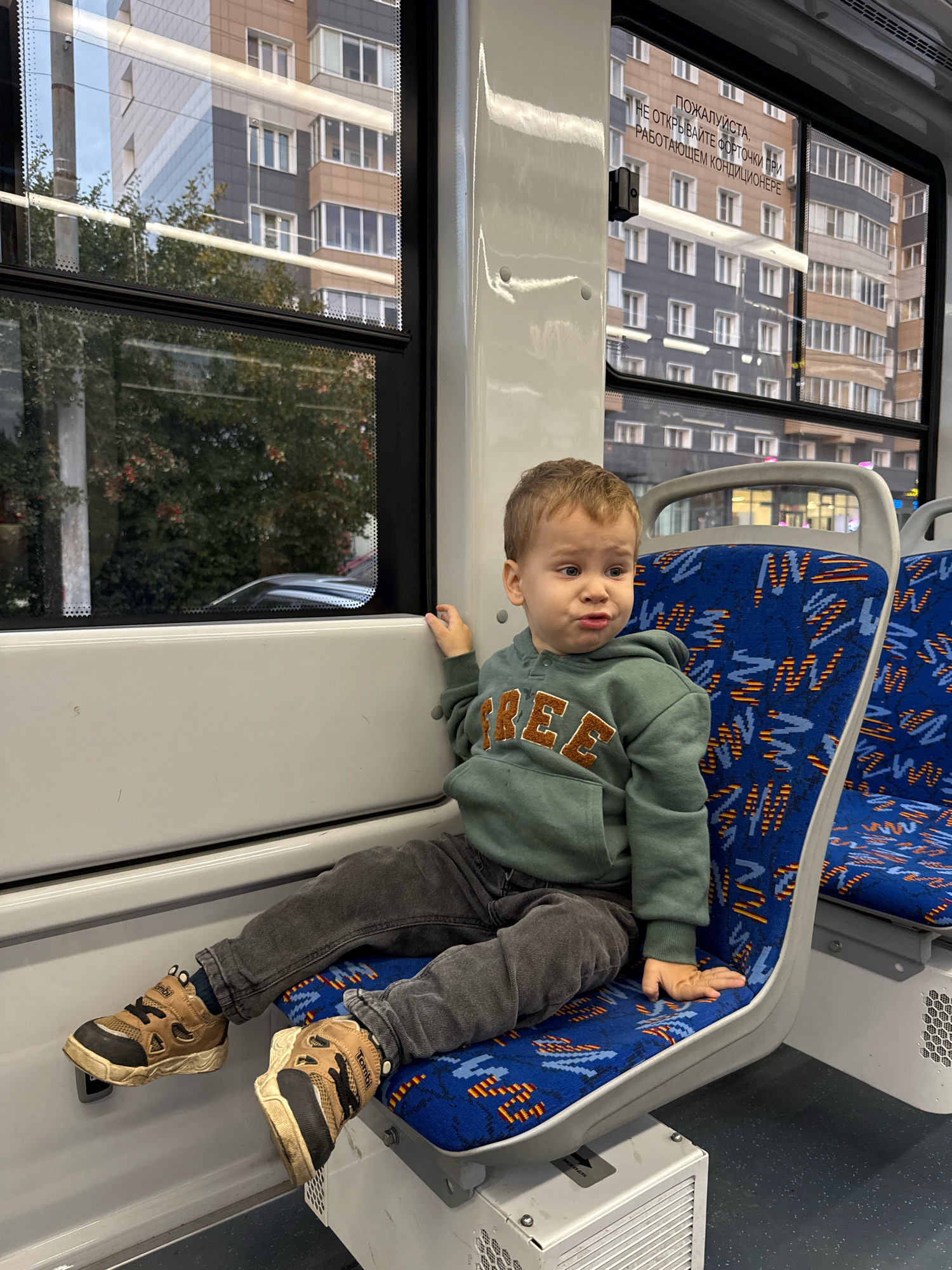 The image shows a young Caucasian boy sitting on a blue and orange patterned seat inside a mode of public transportation, likely a bus or tram. He is wearing a green hooded sweatshirt with the word "FREE" in brown letters across the chest, dark grey pants, and brown and black sneakers. The boy is looking to his right with a slightly displeased expression. His right hand rests on the seat beside him. Through the window to his left, a building with multiple floors and windows is visible, along with trees. The interior of the vehicle is visible, including other seats and white paneling. A sign with Russian text is visible on the window. The environment appears to be urban, consistent with a city setting such as Rusanovo, Russia.