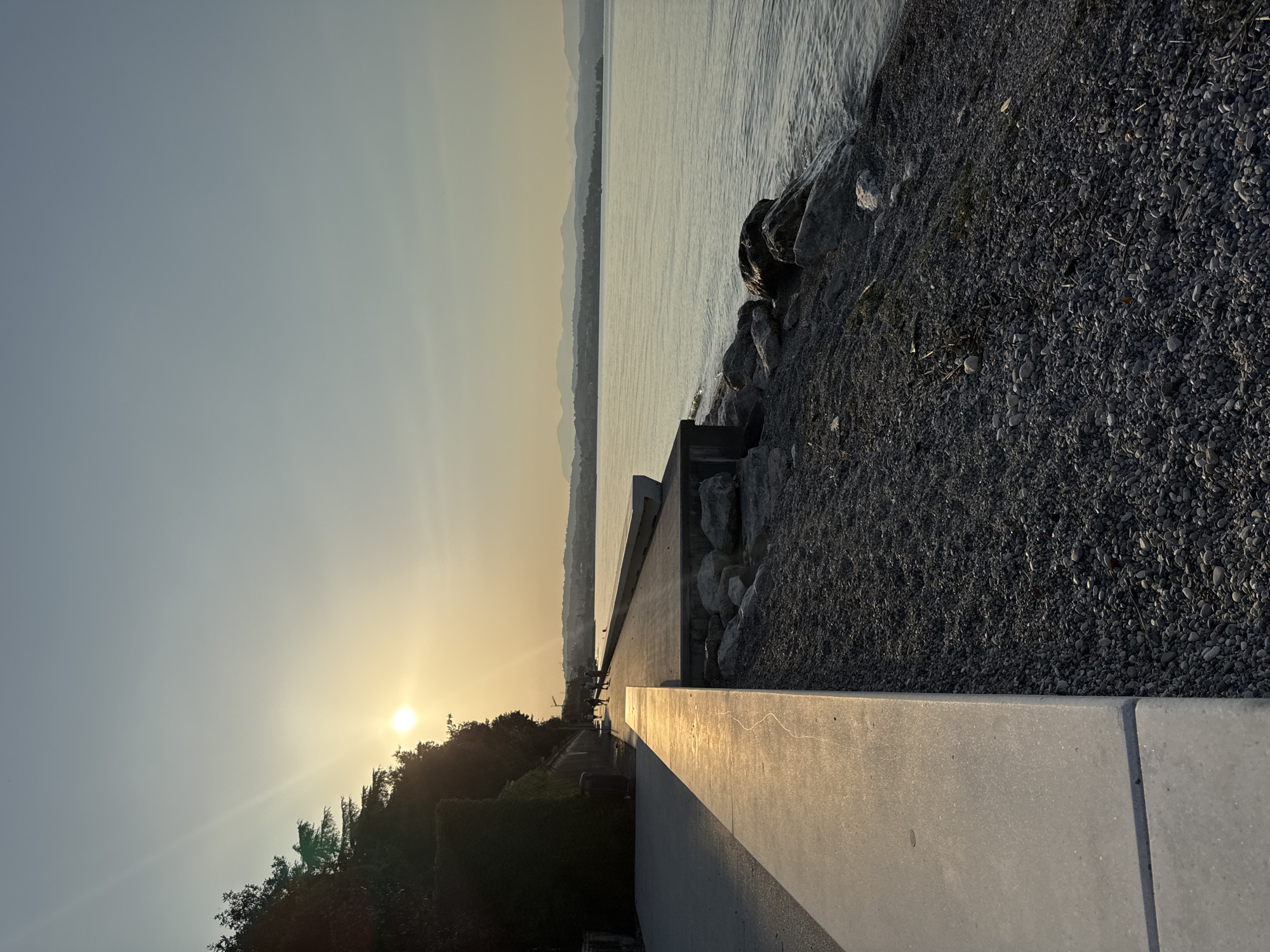The image depicts a serene sunset scene by a lake, likely Desenzano del Garda, Italy. The setting is a paved walkway or promenade adjacent to a rocky shoreline and a calm body of water. Several people are seated or standing on the walkway, enjoying the view. The sun, low on the horizon, casts a warm, golden light, creating a tranquil and contemplative atmosphere. The sky transitions from a clear, pale blue near the sun to a hazy, lighter tone. Distant mountains are faintly visible across the water. The foreground features a concrete structure, possibly a retaining wall, leading to a gravelly area filled with stones and a few scattered twigs. The overall impression is one of peacefulness and the appreciation of natural beauty at dusk.