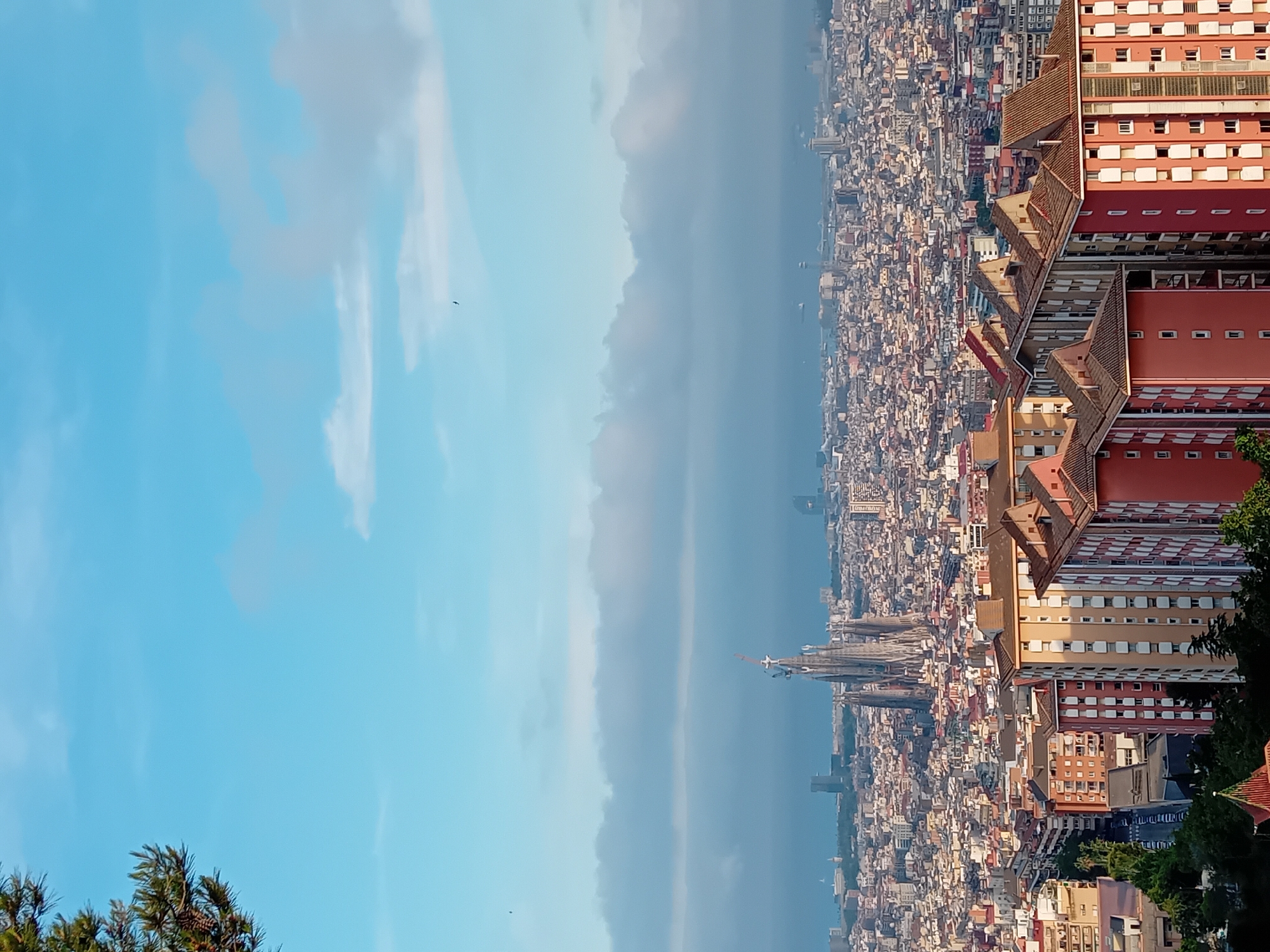 The image shows an elevated panoramic view of Barcelona, Spain. The Sagrada Familia basilica is visible in the middle ground, surrounded by a dense urban landscape. Various buildings, from residential to commercial structures, form a detailed tapestry of the city. Tall, modern buildings stand out among the older construction. In the foreground, a tall red building with multiple windows and rooftops is visible, indicating that the picture was taken from a high vantage point within or near a large structure. The sky is partly clear with some clouds. A small bird is visible in the sky.