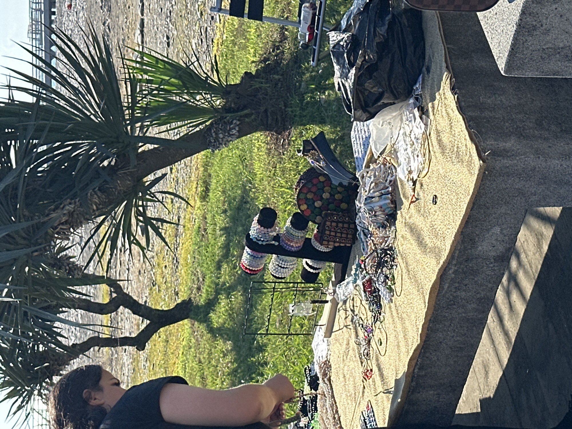 The image depicts an outdoor scene at a market or stall, possibly near a waterfront given the distant view of a pier and beach-like terrain. The foreground features a table covered with a patterned cloth, displaying a variety of handmade bracelets and necklaces. A person, partially visible on the left, appears to be tending to the stall, possibly arranging or selling the items. In the background, palm trees are prominent, suggesting a warm climate. The overall setting is casual and appears to be a public area. The location is identified as Batumi, Georgia.