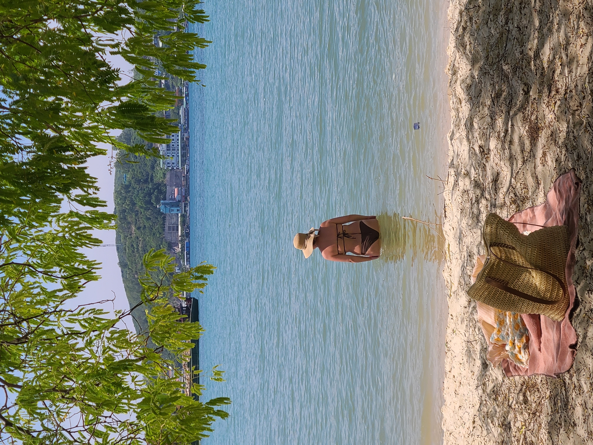 A woman wearing a straw hat and a brown bikini stands in the shallow, turquoise water of the sea. Her back is to the camera, and she is facing a distant cityscape on the shore, which is backed by a green, hilly landscape. In the foreground, a pink towel is spread on a sandy beach, with a woven straw bag and some blankets resting on it. Lush green leaves of a tree hang down from the top of the frame, creating a natural canopy. The scene appears to be a serene beach day in Nha Trang, Vietnam, a coastal city known for its beautiful beaches and vibrant urban backdrop.