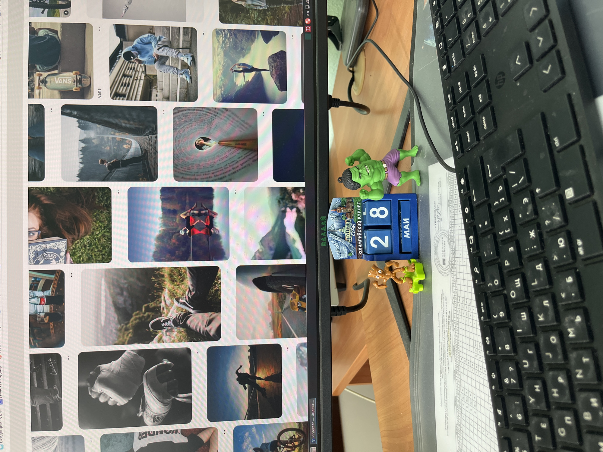 The image captures a desk cluttered with various items, including a computer monitor displaying a Pinterest page with a collection of photos. To the right of the monitor, on the desk, are a black HP keyboard, a sheet of paper with text, and two small figurines: a green Hulk and a brown Groot. A blue calendar cube displays the date "28 MAY" and mentions "SOCHI, Olympic resort." A "ВАЛДАЙ" banner and the text "Олимпийский Курорт" suggest a connection to the Russian city of Sochi. The text on the sheet of paper seems to be in Russian, containing university-related information.