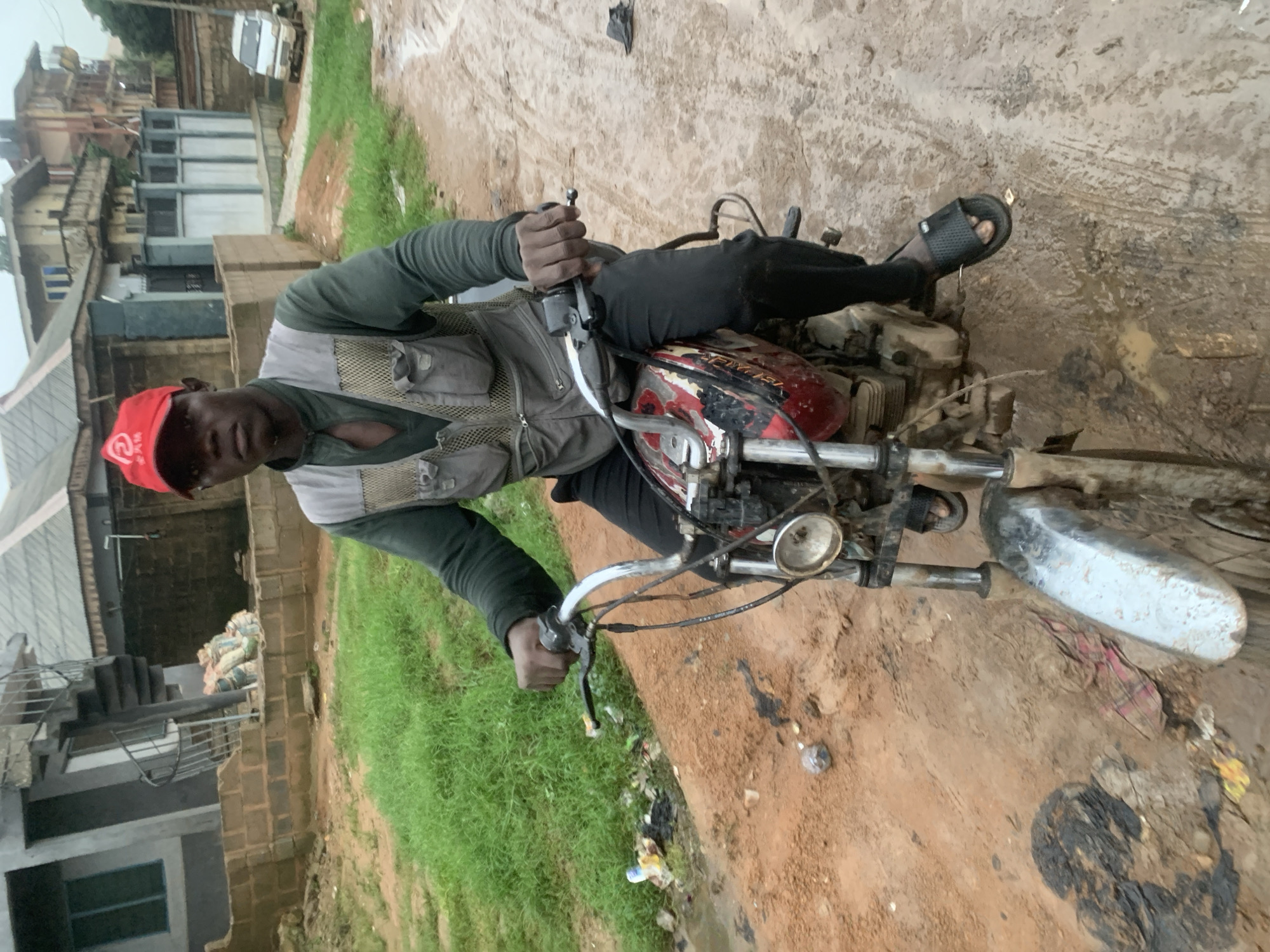In Shagamu, Nigeria, a man wearing a red cap and a gray vest over a dark long-sleeved shirt rides a red and black motorcycle on a dirt road. The road is muddy and appears to be wet, with puddles of water and scattered debris, including plastic bags and what looks like fabric. The man is holding onto the handlebars, and his feet are resting on the footpegs. Behind him, residential buildings with varying architectural styles are visible, some appearing to be under construction. There is also a white van parked in the background. The overall environment suggests an unpaved, somewhat neglected urban area.