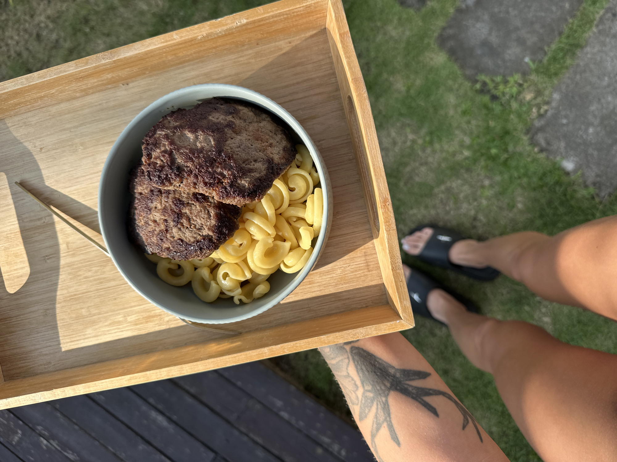 A person is holding a wooden serving tray outdoors in North Kuta, Indonesia. On the tray is a light grey bowl containing cooked pasta and two hamburger patties. The person has a tattoo on their arm, is wearing black slides, and is standing on grass near grey stone tiles and dark wood flooring. The scene is lit by sunlight.