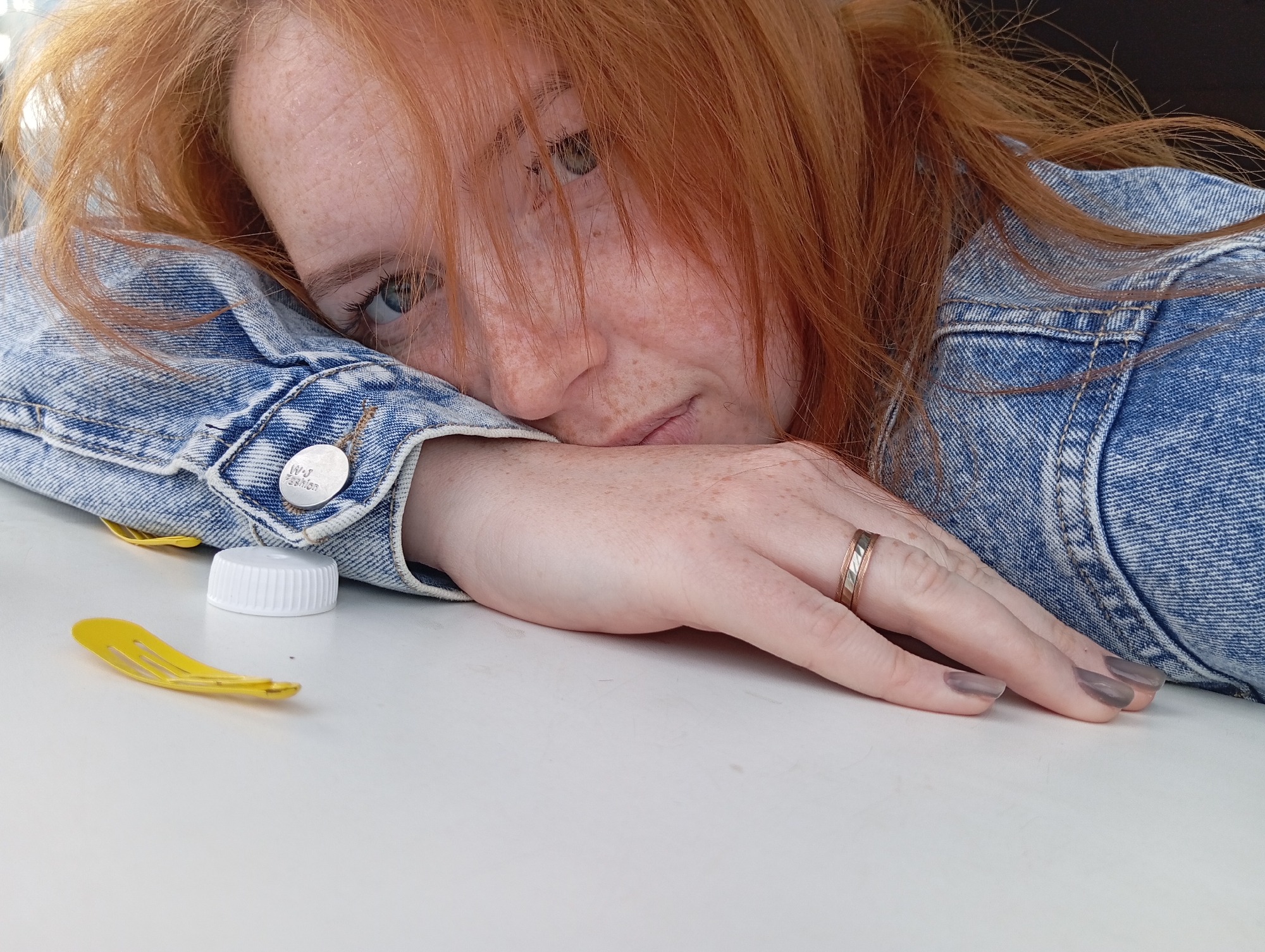 A close-up shot depicts a fair-skinned, freckled, light-eyed person with red hair resting their head on their denim-clad arm. The person's hand, adorned with a simple gold band, lies palm down on a white surface. A yellow plastic spork and a white plastic bottle cap are positioned near their hand. The background is a blurry mix of dark and light tones. There is no data to suggest that this image was taken in Yurgamysh, Russia.
