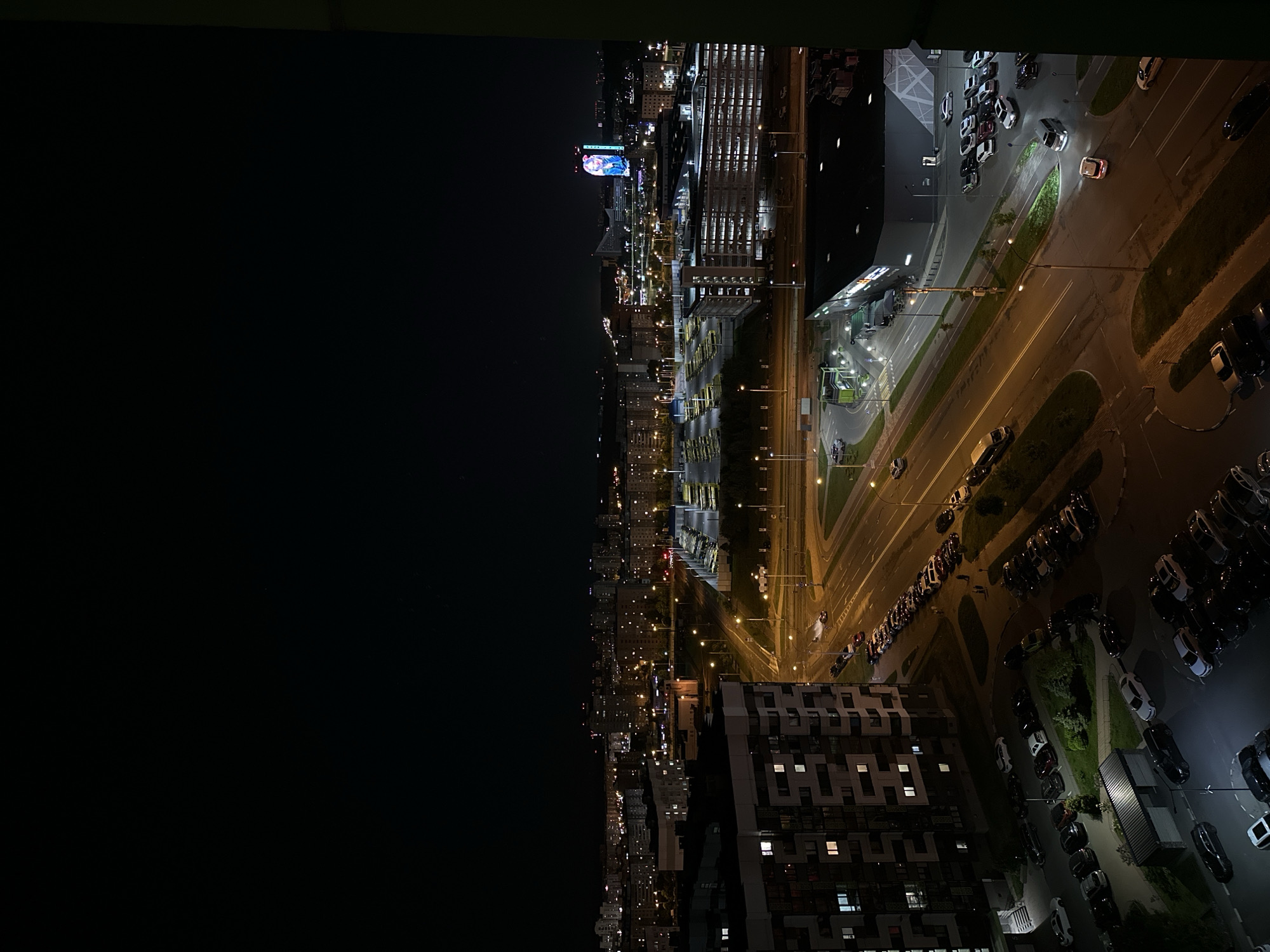 A nighttime aerial view of a portion of Бараўлянскі сельскі Савет, Belarus. The left half of the image is dominated by a black sky. The right half depicts a lighted urban scene. At the center is a road with street lights, with cars driving along it. Adjacent to the road is a parking lot filled with numerous parked cars. Beyond the road are buildings, including a large structure with rows of buses parked in front. Further in the distance are more buildings and illuminated billboards. At the bottom, the corner of a multi-story building with lit windows is visible.