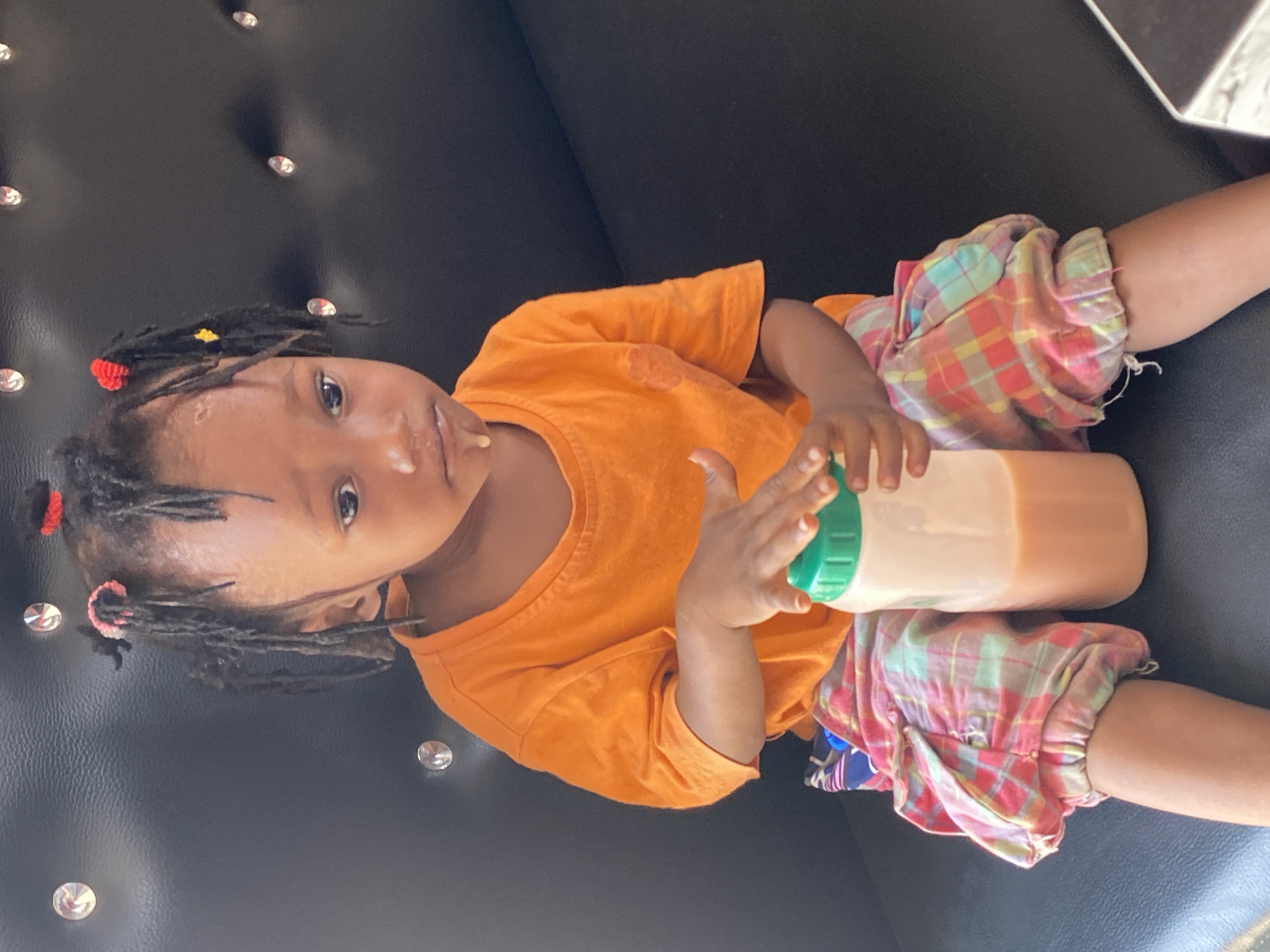 A young child, likely a toddler, is shown lying on a black, tufted piece of furniture adorned with shiny, silver-colored studs. The child has dark skin, wide, dark eyes looking directly at the camera, and short dark hair styled in dreadlocks or twists, accented with small red and yellow hair ties. There is residue around the child's mouth and chin, possibly from a drink or food, and a small mark is visible on the forehead.
The child is dressed in a bright orange short-sleeved t-shirt and colorful plaid shorts featuring a mix of red, pink, green, yellow, and blue squares. They are holding a beige-colored bottle with a bright green cap, which contains a light brown liquid. The setting appears to be indoors with adequate lighting, suggesting it is daytime. The child's expression is neutral and attentive. This scene is located in Adankolo, Nigeria. There is no visible text in the image.