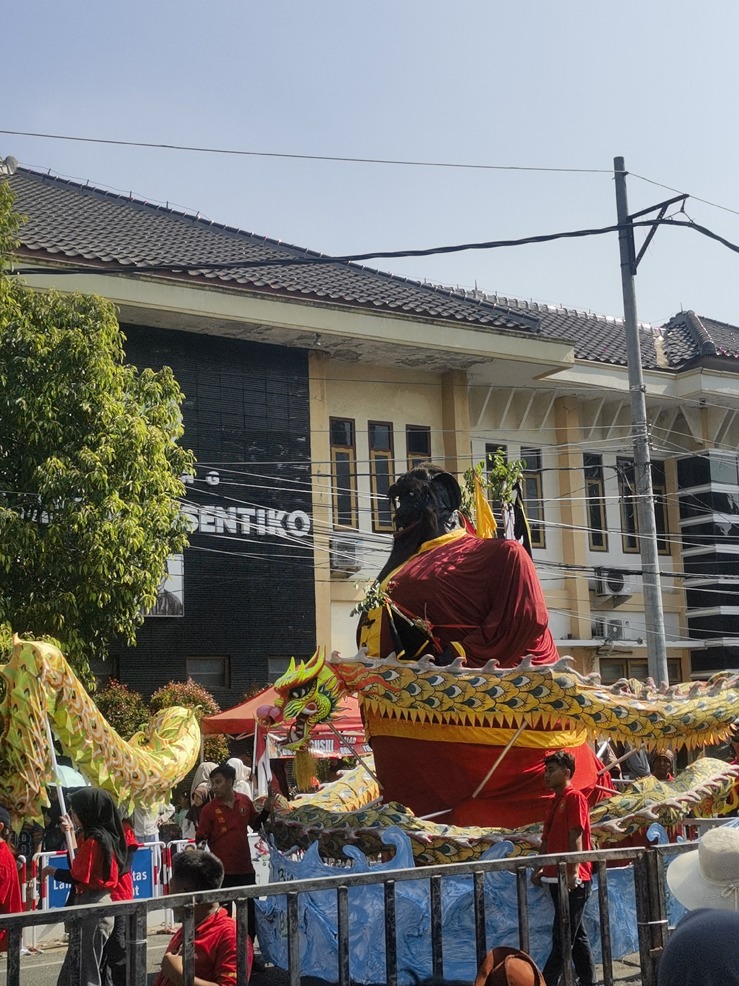 In an outdoor daytime scene in the city of Unknown, Unknown, a large-scale parade float is the central focus. The float features a giant, dark-faced effigy wearing a vibrant red robe. This effigy is adorned with a golden, dragon-like structure that wraps around it, decorated with yellow scales and red accents. Several people are visible around the float, some dressed in red, participating in the parade. In the background, a building with a dark facade and windows can be seen, with a large sign that reads "SENTIKO". A lush green tree stands to the left of the building, and a utility pole with wires is positioned to the right. The sky above is clear and blue.