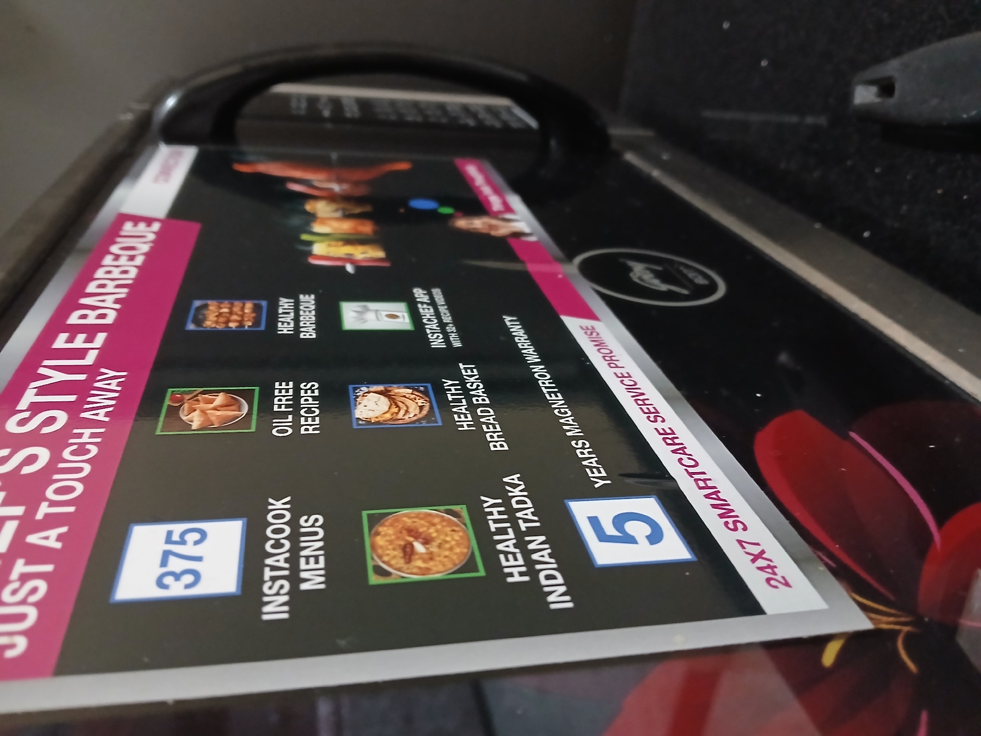 A close-up, slightly angled shot shows the surface of a cooking appliance, likely a microwave oven, featuring a promotional graphic with a pink vertical bar on the left. The text on the graphic includes "JUST A TOUCH AWAY," "375 INSTACOOK MENUS," "OIL-FREE RECIPES," "HEALTHY INDIAN TADKA," "5 YEARS MAGNETRON WARRANTY," and "24X7 SMARTCARE SERVICE PROMISE."
There are various images corresponding to "HEALTHY BARBEQUE," "INSTACHEF APP," and "HEALTHY BREAD BASKET." The context implies a domestic setting. The image was likely taken in Bengaluru, India, given the regional cuisine displayed in the imagery.