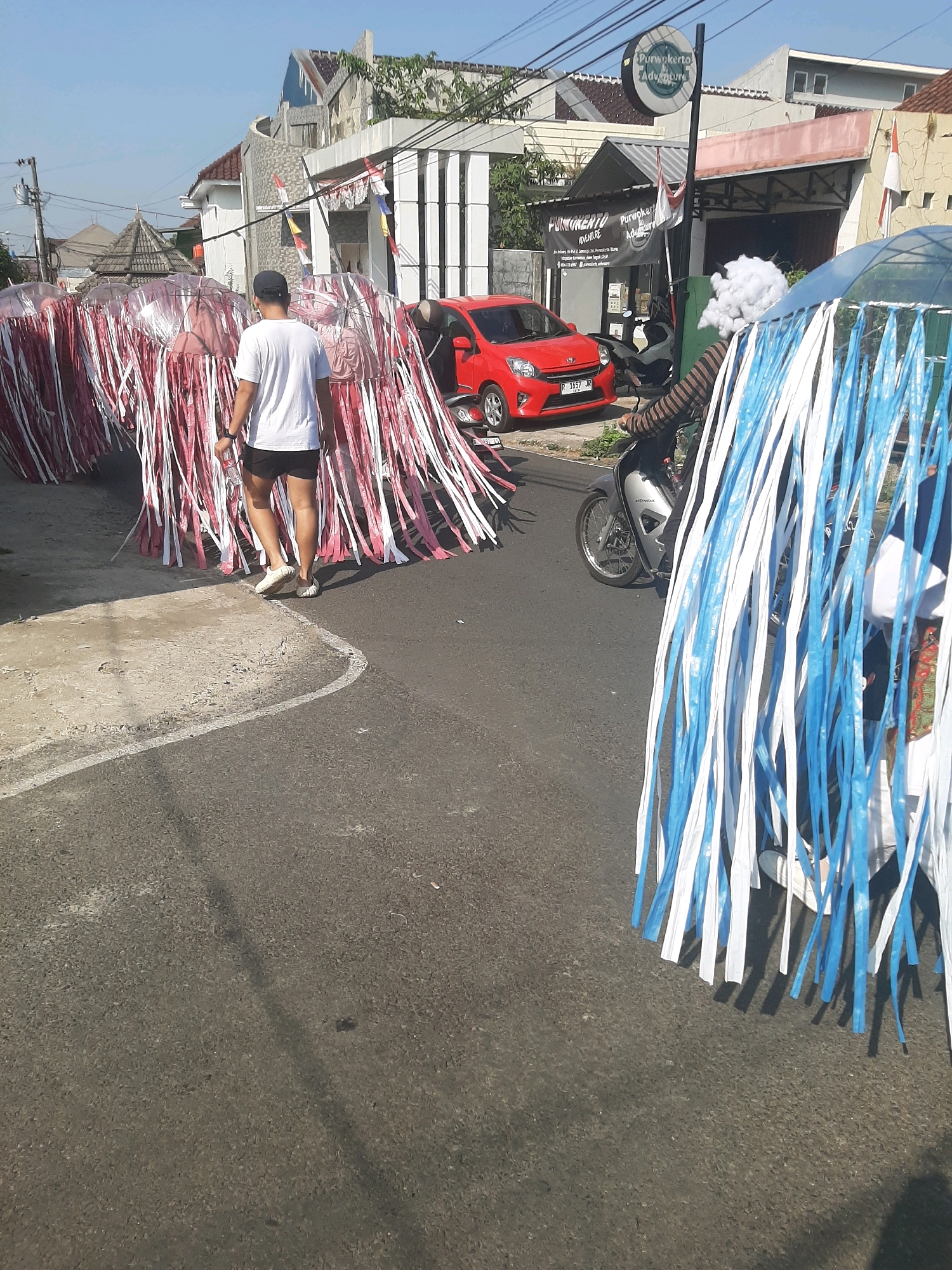 In Sumampir, Indonesia, a man wearing a white t-shirt and black shorts walks on a paved road. He holds a water bottle and is nearing a collection of large, elaborate decorations made of red and white streamers cascading from clear umbrella-like structures. To his right, a motorcycle is parked. Further to the right, another similar decoration, this one with blue and white streamers, is visible. A person on the motorcycle wears a striped shirt and has a cloud-like white object on their head. In the background, buildings and houses line the street under a clear blue sky. A red car is also parked on the side of the road.