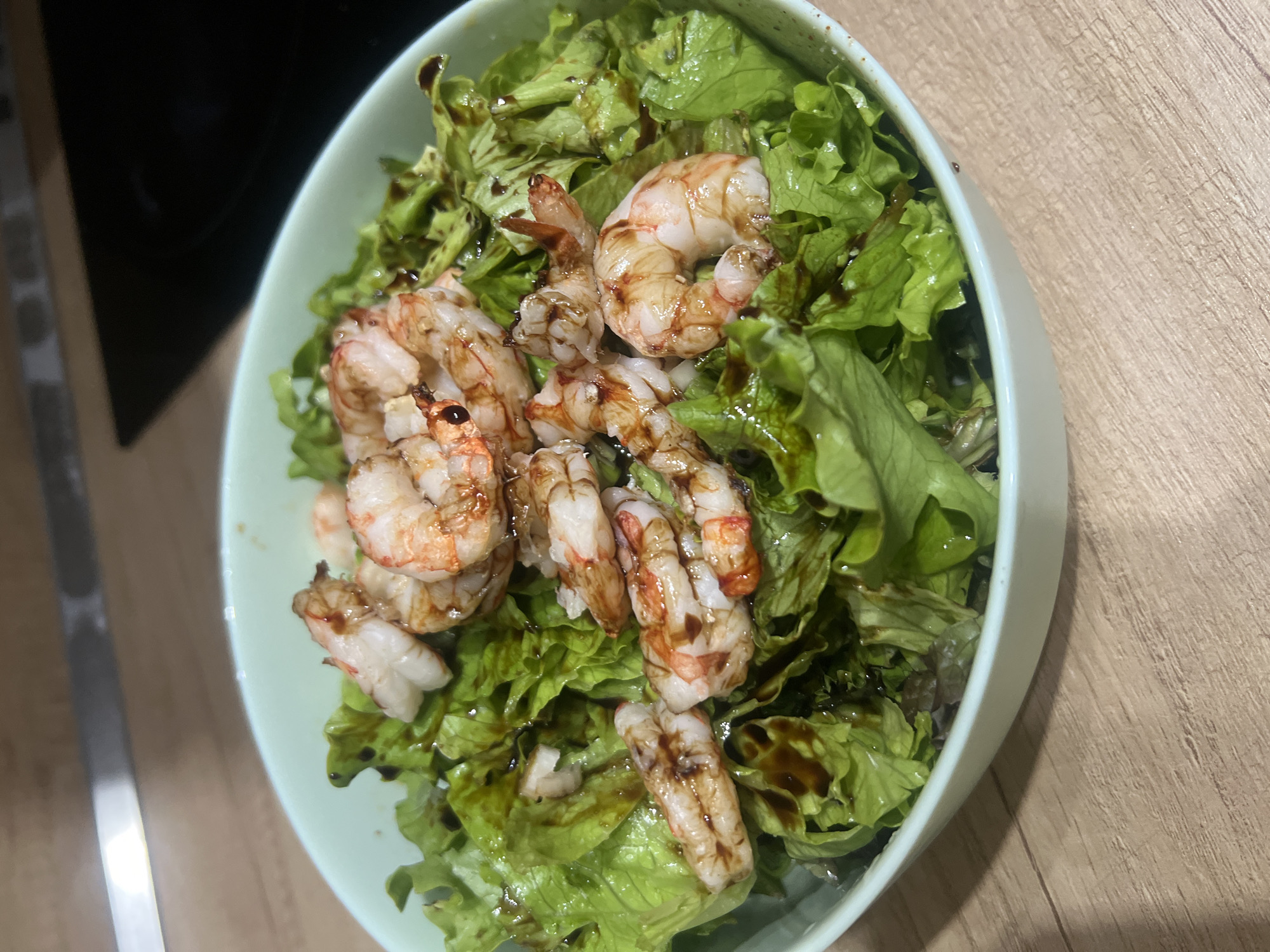 The image shows a bowl of salad with shrimp, likely on a wooden countertop. The salad consists of green lettuce leaves, and the shrimp are arranged on top, drizzled with a dark sauce, possibly balsamic glaze. The bowl appears to be light green or blue. The location is likely a private residence. Nothing suggests it is in Plovdiv, Bulgaria; there are no contextual elements linking the photo to that location.